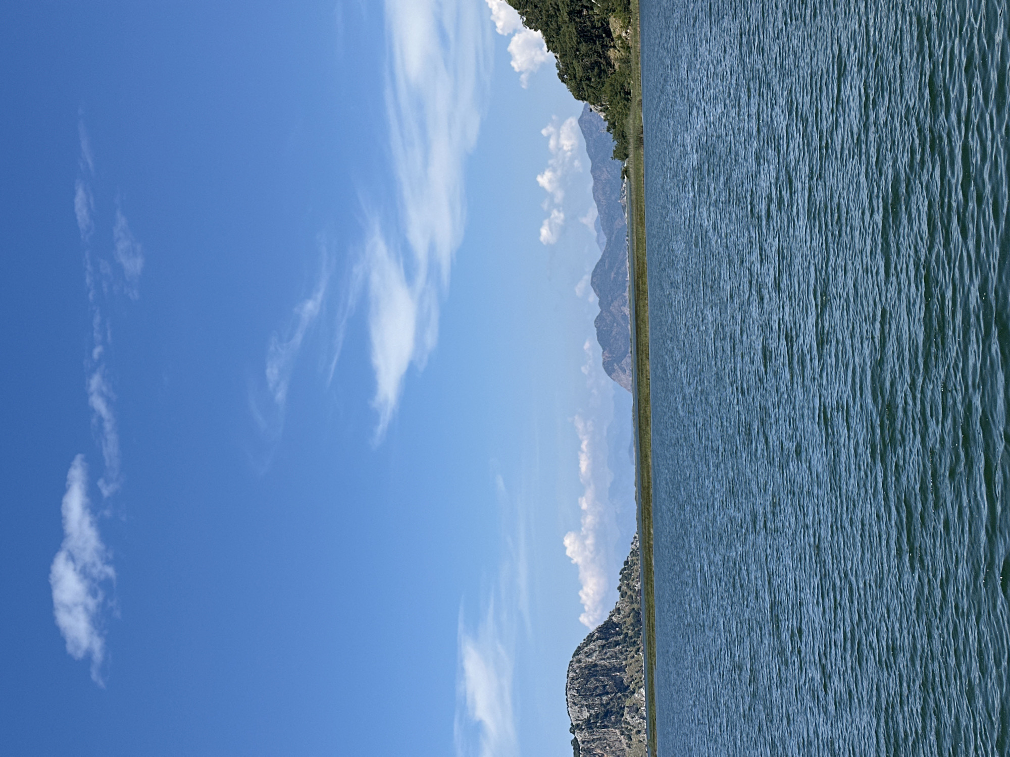 The image shows a landscape with a body of water, mountains, and sky. The water is on the right side of the image and shows signs of ripples or small waves. A thin strip of land separates the water from the mountains in the distance. The mountains appear to be rocky and sparsely vegetated in some areas. The sky on the left side is a clear blue with scattered white clouds.
Based on the location provided, this scene could potentially be from Köyceğiz Lake in Ortaca, Turkey, given the presence of a large lake surrounded by mountains in that region.