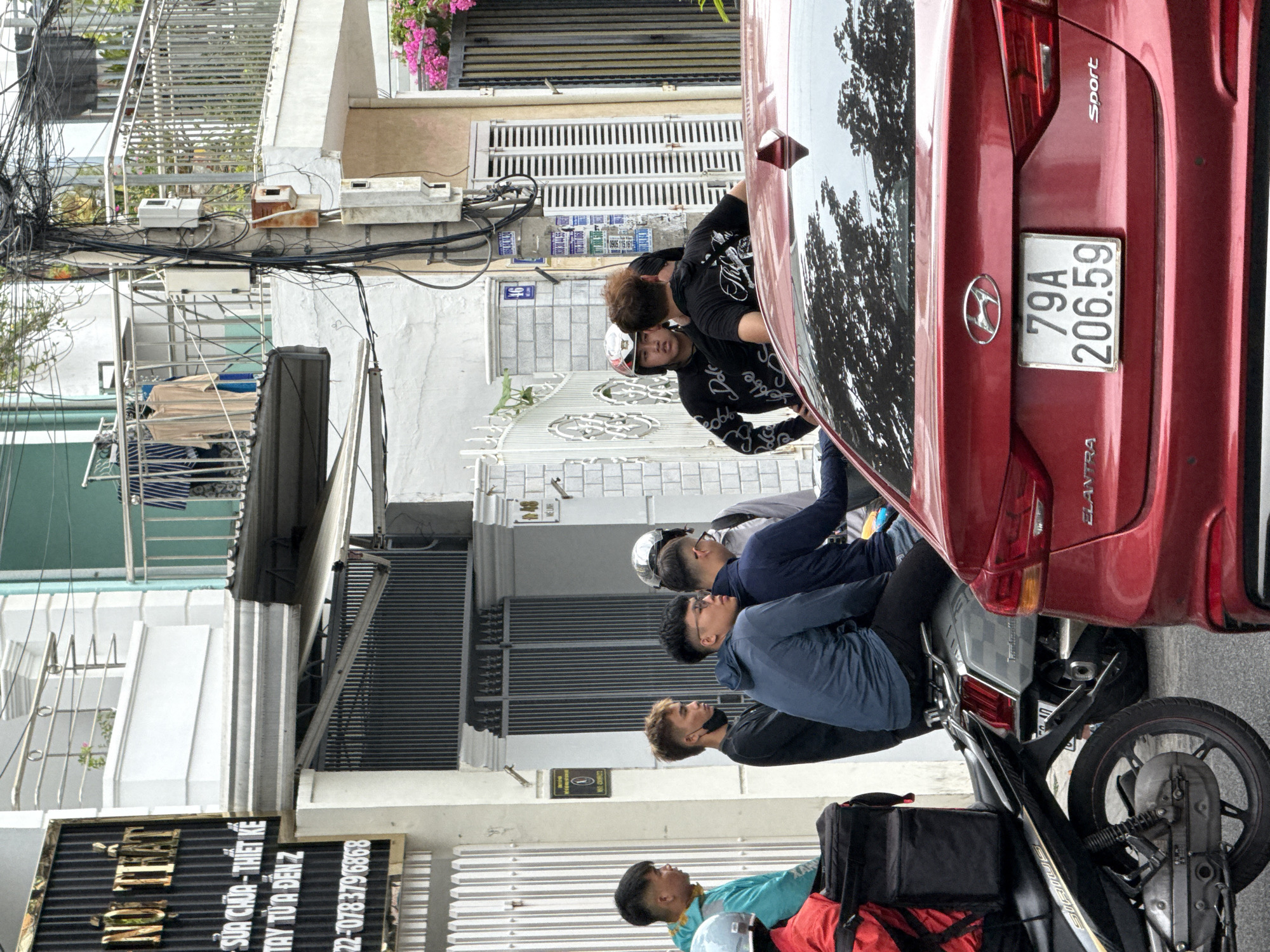 The image depicts a street scene in Nha Trang, Vietnam. In the foreground, a red Hyundai Elantra is parked, its rear visible with a license plate reading "79A 206.59". To the left of the car, a motorcycle with a black delivery box and red accents is parked. Several individuals are gathered on the sidewalk. From left to right, a person in a teal jacket is visible, followed by a person in a black shirt, and then two individuals in blue and navy jackets, respectively, sitting on a motorcycle. Further back, a group of men are standing and conversing, some wearing black t-shirts and one wearing a helmet. Behind them, buildings with balconies and various electrical wires are visible. The environment appears to be an urban setting with typical street elements like signage, fences, and vegetation.