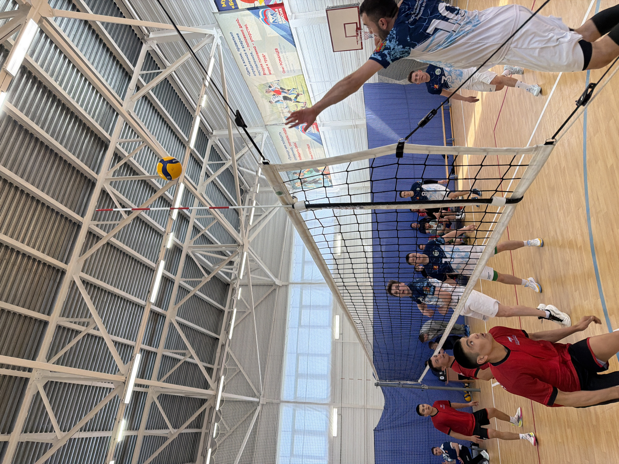 This is an indoor volleyball game. One player wearing a blue and white jersey is jumping high to hit the ball over the net, while another player wearing a red jersey is looking up at the ball, ready to receive it. Several other players in both blue and red jerseys are present on the court. The game appears to be taking place in a gymnasium, indicated by the high ceiling with metal support structures and lighting, as well as a banner on the wall with text in Russian. Some of the readable text on the banner includes: "Bolshaya Murta" and "Russia," indicating that the location is in the city of Bolshaya Murta, Russia. There's also a banner with the words "ЕДИНАЯ РОССИЯ" which could be translated as "United Russia" in English.