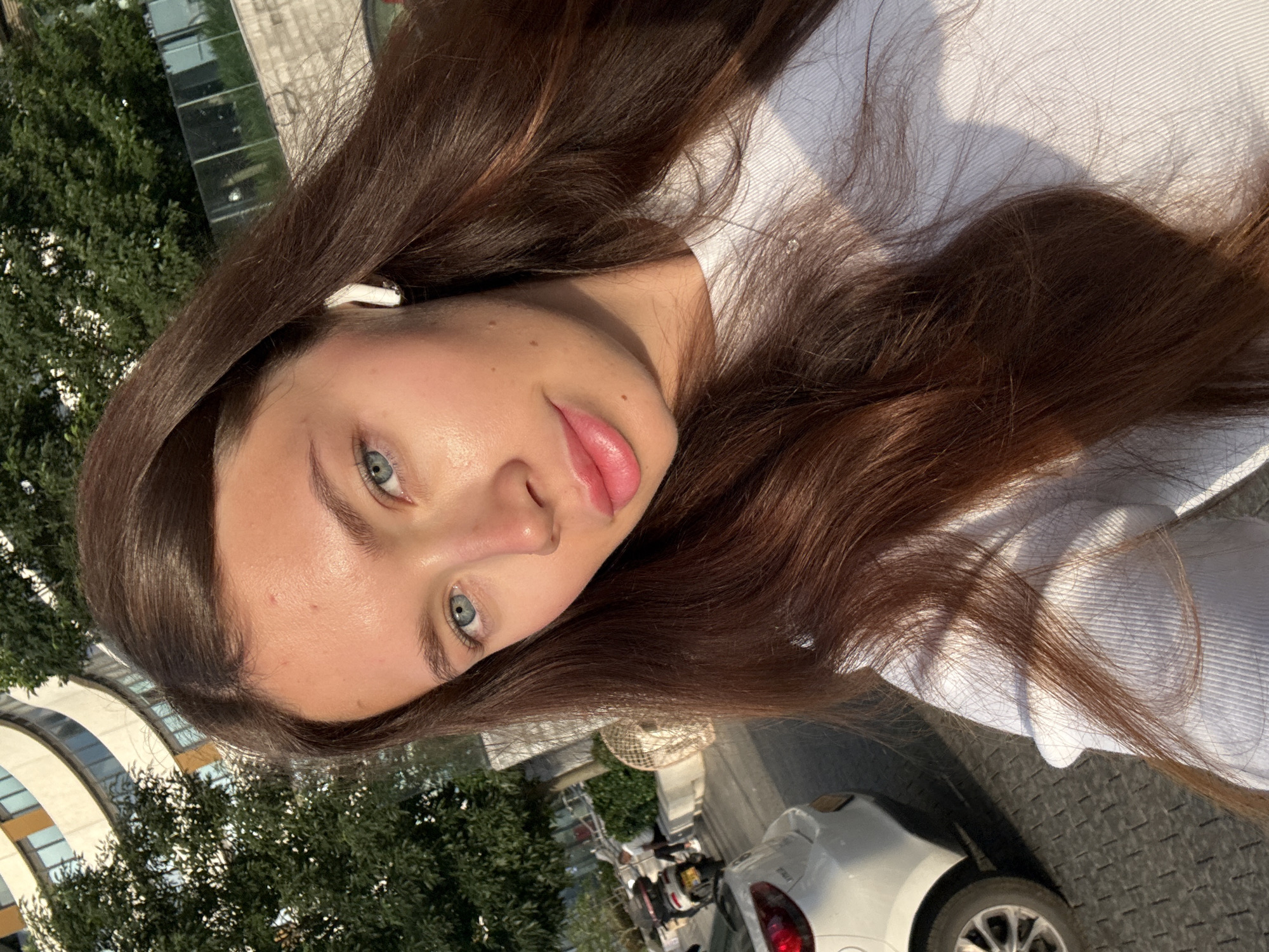 A young woman with long brown hair and blue eyes is pictured in a selfie. She is wearing a white shirt and has a wireless earbud in her left ear. The background shows an urban street scene with trees, buildings, and a white car parked on the side of the road. The location is likely Tel Aviv, Israel.
