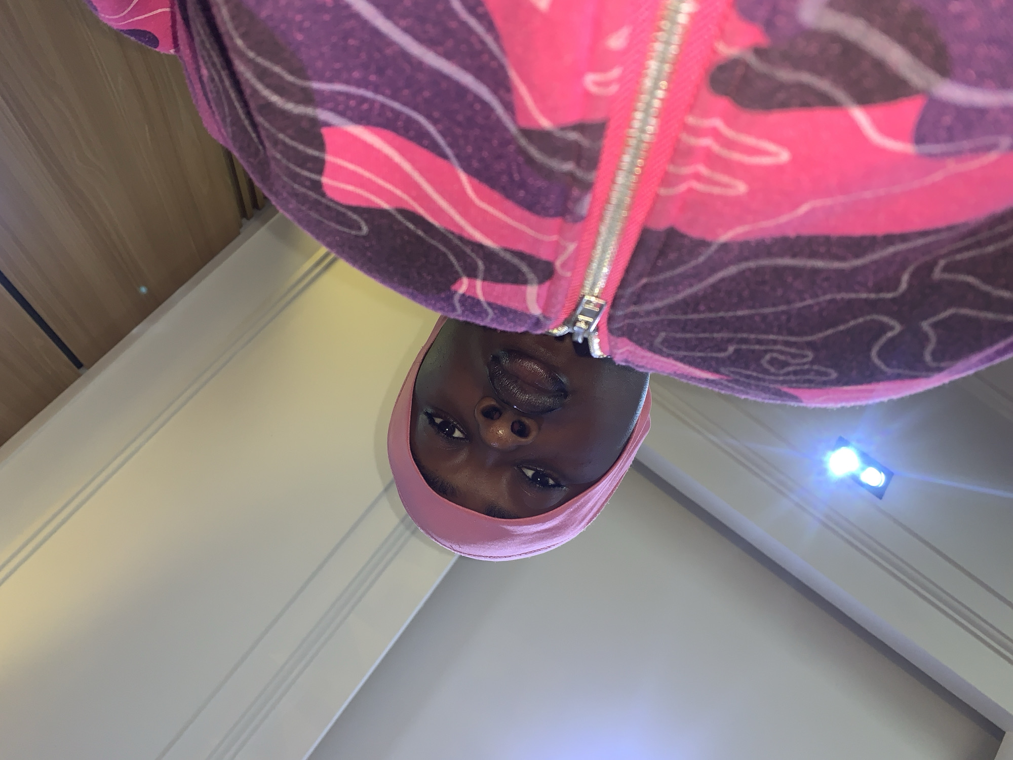 The image shows a close-up, low-angle shot of a person wearing a pink bonnet and a purple and pink patterned zip-up hoodie. The person's face is turned towards the camera, with their lips slightly parted. The background consists of a white ceiling with light fixtures, and a wooden element at the top of the frame. The scene appears to be indoors, possibly in a residential setting. No specific location in Gura Top, Nigeria can be determined from the image itself.