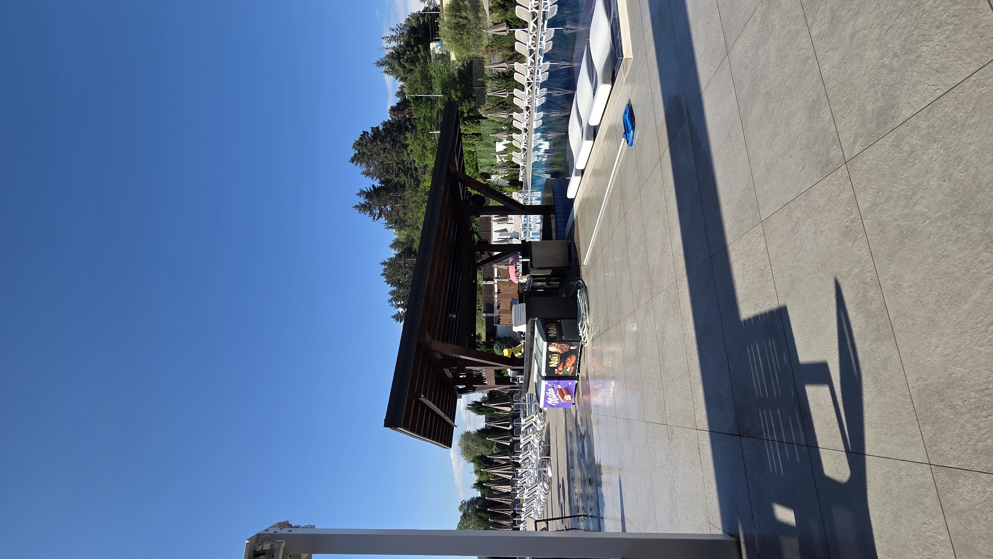 The image shows an outdoor swimming pool area on a sunny day, with a clear blue sky. In the foreground, a tiled patio area is visible, with a long shadow cast by a covered structure. This structure appears to be a bar or a seating area with a dark wooden roof supported by pillars. Underneath the roof, there are some counters and what look like refrigerators, one displaying a "Milka" logo and another with "Nuii" branding. To the left of the structure, numerous lounge chairs with open umbrellas are neatly arranged. To the right, the swimming pool is visible, reflecting the sky and surrounding trees. The pool has white submerged loungers. In the background, a dense forest covers a hillside, with a few buildings peeking through the trees. The overall scene suggests a resort or a recreational facility in a natural setting, likely in Bania, Bulgaria, given the context. There are no people clearly visible in the image.