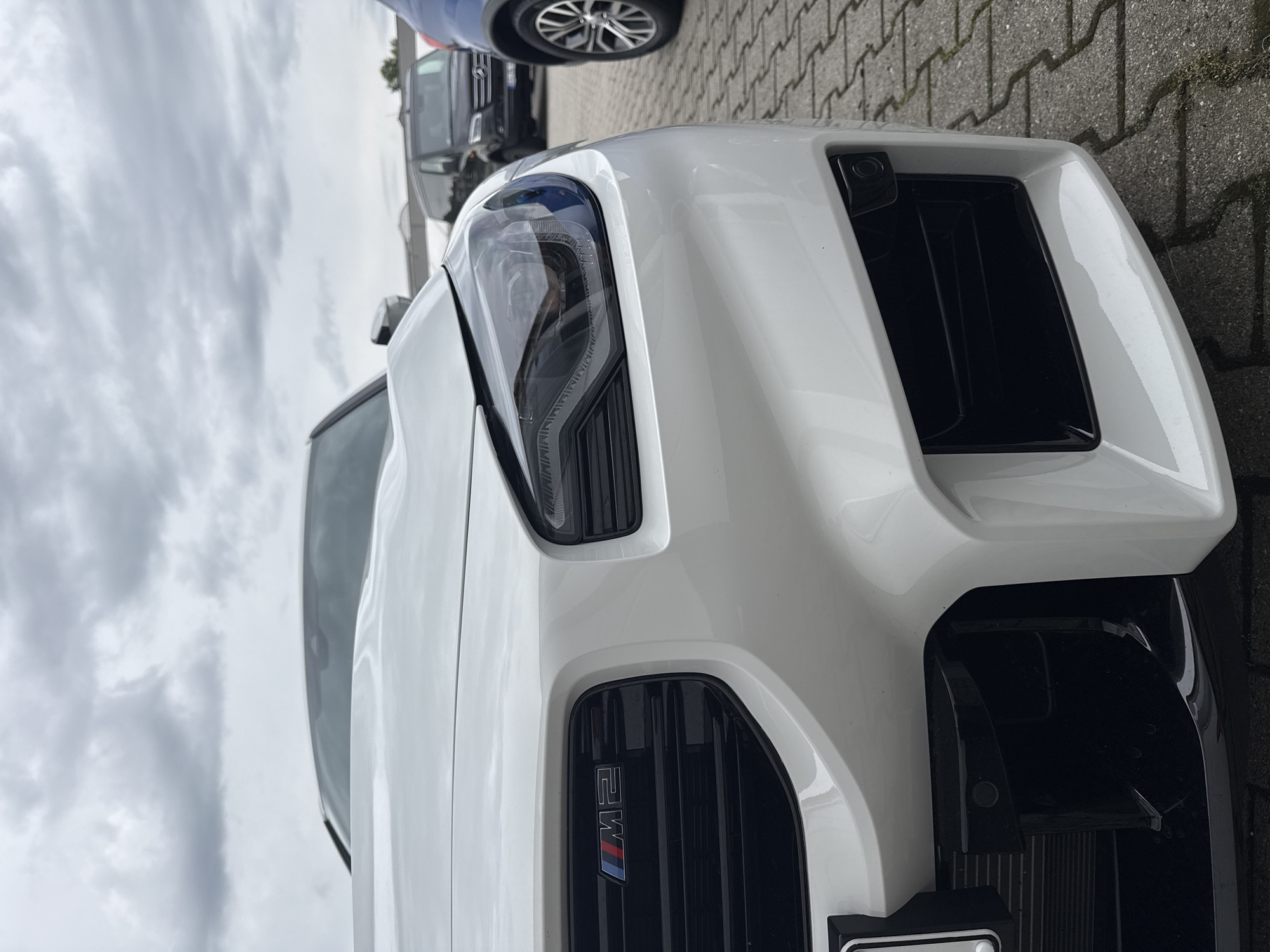 This media file shows a close-up, low-angle, vertical shot of the front of a white BMW M2 parked on a paved area with a cobblestone-like pattern. The car's headlight and grill are prominently featured. The grill has the "M2" badge with the iconic M colors. To the left of the car, a portion of a dark-colored van is visible, and behind it, the wheel of a blue vehicle can be seen. The sky is overcast with dramatic clouds.
The scene appears to be outdoors, possibly a dealership or parking lot, given the presence of multiple vehicles and the paved ground. The time of day is likely daytime, but the cloudy sky suggests it might be overcast or nearing evening. There are no people visible in the frame. The focus is entirely on the car's front fascia and the surrounding environment. The image is sharp and detailed, highlighting the car's design elements and the texture of the ground. No specific activity is depicted, suggesting the car is stationary.
The location context provided is Wettstetten, Germany.