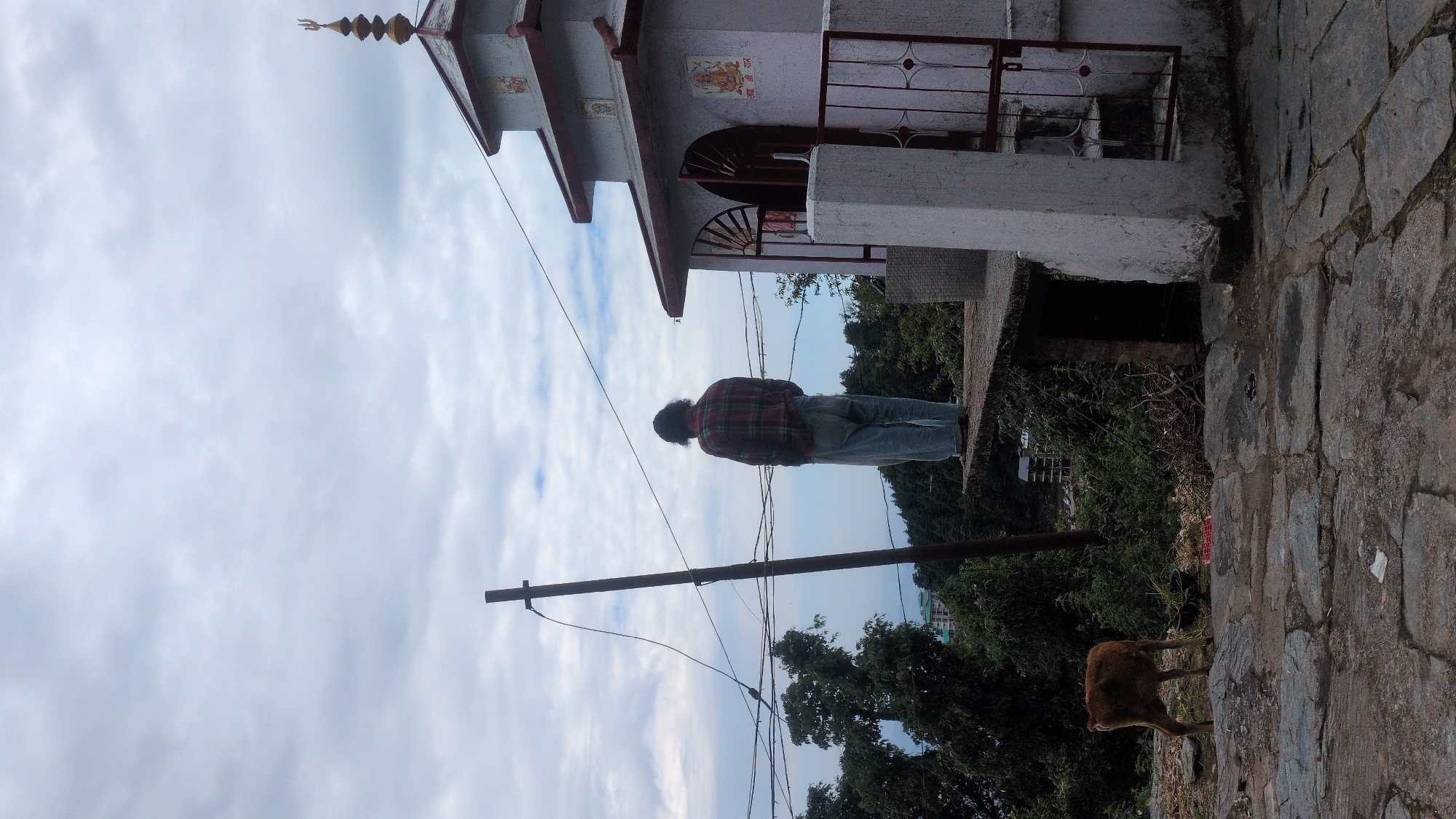 An outdoor scene in Haini, India, captured on an overcast day, features a person, a small temple, and an animal, set against a cloudy sky.
In the center of the frame, a person stands with their back to the camera, looking towards the right side of the image. They have dark, curly hair and are wearing a red and black plaid shirt over blue jeans.
To the left, a small, white temple or shrine is partially visible. It has a multi-tiered, sloped roof topped with a golden finial resembling a trishul (a trident, a significant Hindu symbol). The temple entrance features an arched doorway with decorative metalwork. On the white wall above the archway, the Hindi text "श्री राम" (Shri Ram) is clearly visible.
A sturdy wooden utility pole with multiple wires spans across the middle ground. The ground in the foreground and extending to the right is paved with irregular, large stone slabs, indicating a rugged path or courtyard.
In the lower right corner, a brown animal, likely a cow or calf, is partially visible, walking or grazing on the stone path. Lush green trees and bushes fill the background and midground, suggesting a natural, possibly hilly, environment. The sky is largely covered with white and grey clouds, indicating an overcast or partly cloudy day. The overall atmosphere appears calm and tranquil.