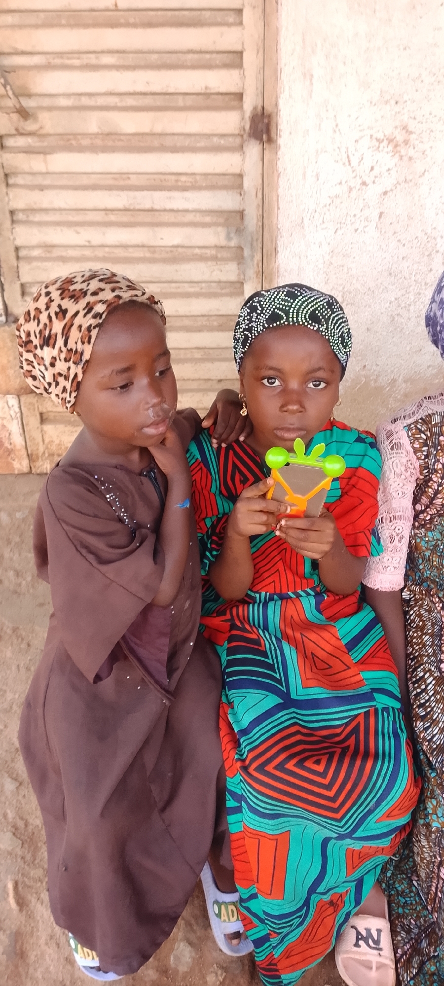 Two young girls sit side-by-side outdoors, possibly in a shaded area. The girl on the left wears a brown robe and a leopard-print headscarf. The girl on the right wears a vibrantly patterned dress with red, blue, and green geometric designs and a black and white patterned headscarf. She holds a small, colorful toy in her hands. Both girls have dark skin and dark hair. Behind them, a wooden structure with horizontal slats is visible, and the background appears to be a textured wall. The ground beneath them is sandy or dusty.