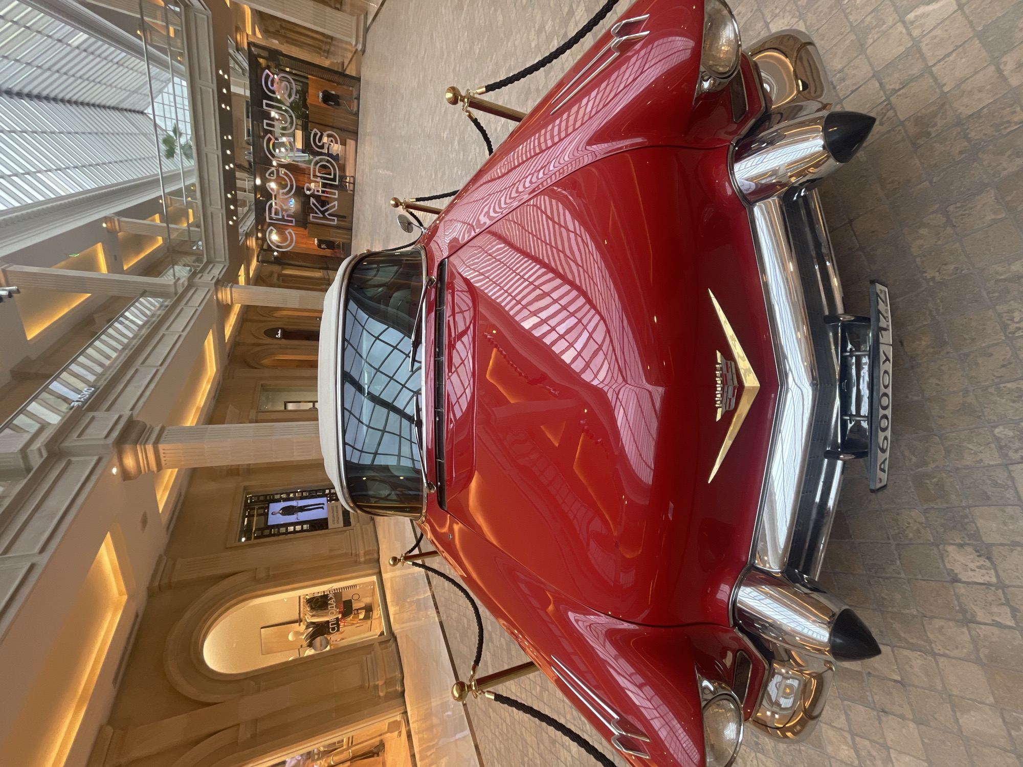 In the atrium of the Crocus City Mall in Khimki, Russia, a vintage bright red convertible Cadillac is prominently displayed. The car is positioned centrally, facing the camera, with its gleaming chrome accents and cream-colored convertible top clearly visible. A black rope barrier, supported by gold stanchions, encircles the front of the vehicle. Behind the car, the mall's interior features ornate architectural details, including tall columns and arched storefronts, illuminated by warm, ambient lighting. The reflection of the glass ceiling and the mall's upper levels can be seen on the car's polished surface. In the background, signage for "CROCUS KIDS" is visible, along with displays in shop windows. The floor is paved with a light-colored tile pattern.
