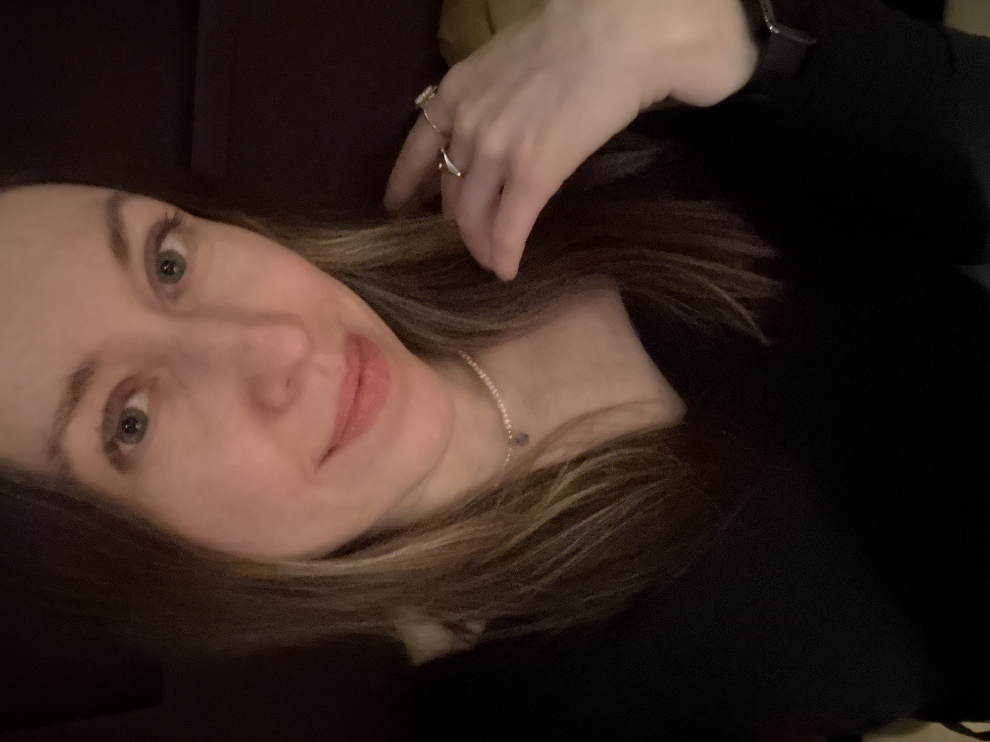 A selfie-style close-up photo captures a fair-skinned woman with light brown hair and green eyes. She wears a black long-sleeved top and a delicate necklace. Two rings adorn her left hand, which is partially visible near her face. A dark-colored surface is visible in the background. The location is not discernible from the image. No details suggest Saint Petersburg, Russia.
