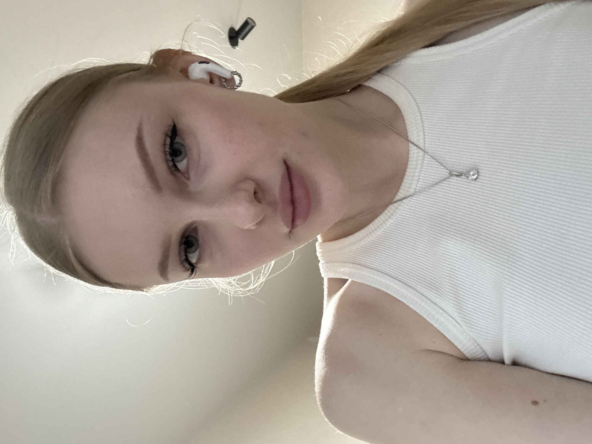 A close-up, downward-angled portrait of a young woman with light blonde hair. She is wearing a white tank top and a delicate silver necklace with a small pendant. She has fair skin and her eyes are green. An earbud is visible in her left ear. The background is a plain, light-colored wall. It is not possible to determine a location in the city of Perm, Russia, from this image.