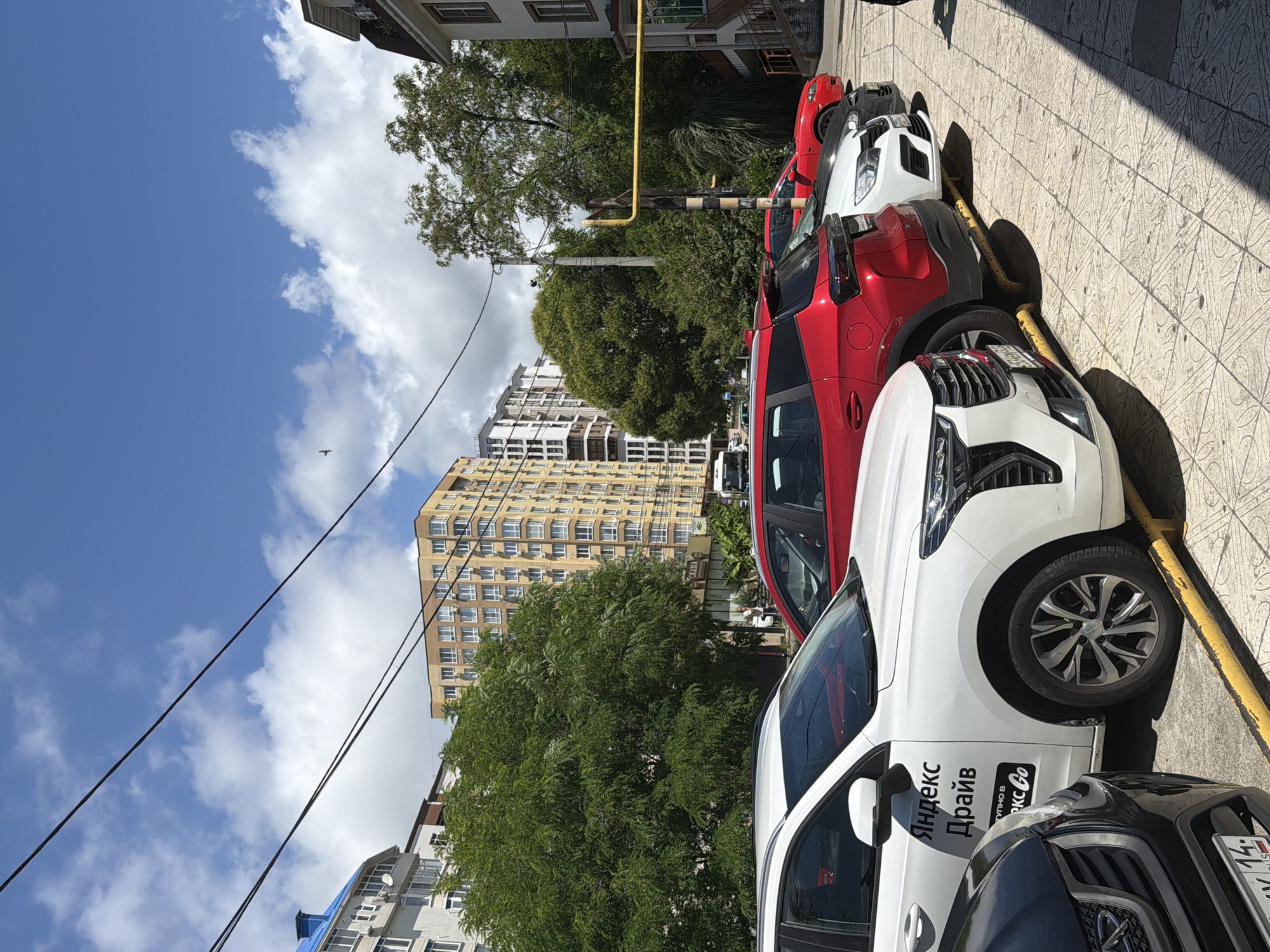 The image depicts a street scene in Sochi, Russia, with several cars parked in a row on a paved area. The foreground features the front and side of a white vehicle with "Яндекс Драйв" (Yandex Drive) branding, indicating it's a ride-sharing car. Behind it, a red SUV is parked, followed by a white car and another red car further down the line. The background reveals a dense urban environment with multi-story buildings of varying architectural styles and colors, including a prominent yellow building. Lush green trees are interspersed between the buildings, suggesting a well-treed city. The sky is blue with scattered white clouds, and power lines are visible overhead. The scene appears to be a typical street in a residential or commercial area.