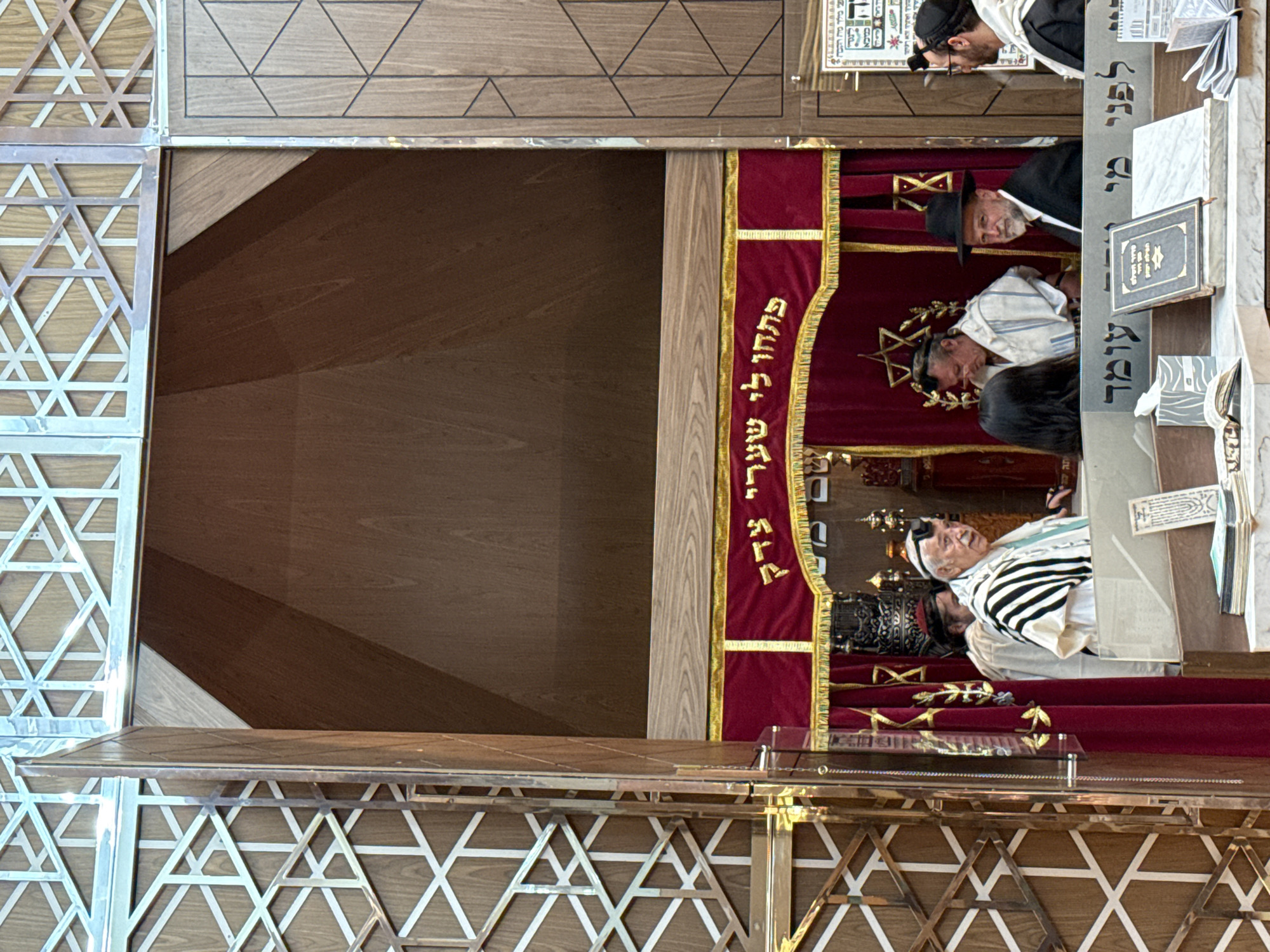 The image shows a section of a synagogue in Mexico City. Three men wearing traditional Jewish attire stand behind a wooden partition adorned with a red velvet curtain with gold embroidery, presumably an Ark housing Torah scrolls. The partition itself is framed by a decorative screen featuring the Star of David. In front of the men and the partition is a reading table with books and papers.
