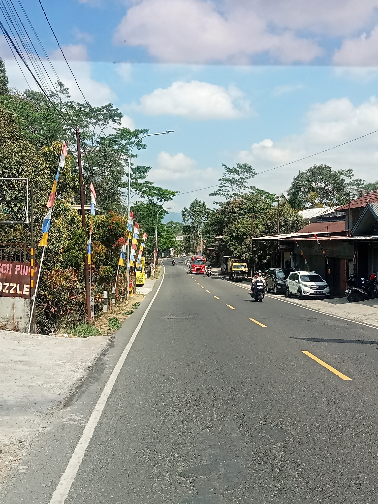 The image depicts a sunny day on a paved road in Selokromo, Indonesia. The road is lined with trees and buildings on the right side. Several small flags, predominantly red, white, blue, and yellow, are planted along the roadside. A white car and several motorcycles are visible on the road and parked near the buildings. A red truck is seen in the distance, traveling away from the viewer. The sky is blue with scattered white clouds. A sign on the left side of the road reads "SCH PUMP OOZZLE". The scene appears to be a street with moderate traffic and a commercial area.