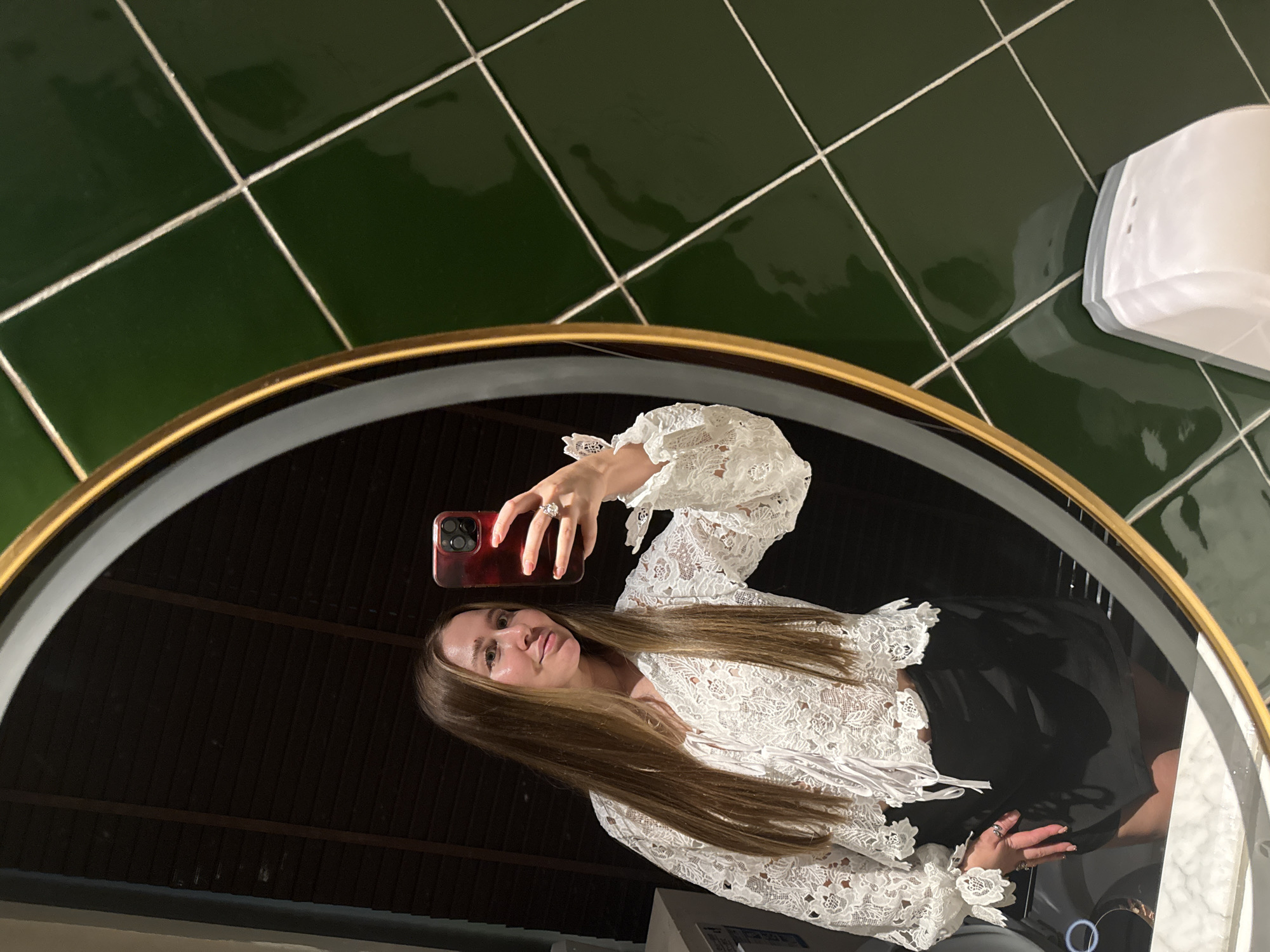 In Yiwu, China, a woman stands in front of a mirror, taking a selfie. She is wearing a white lace blouse with voluminous sleeves and a black skirt. The mirror reflects her and part of a room with dark blinds. To the right of the mirror, green square tiles with white grout cover the wall. A white dispenser is visible on the tiled surface. The woman holds a red-cased smartphone to take the picture.