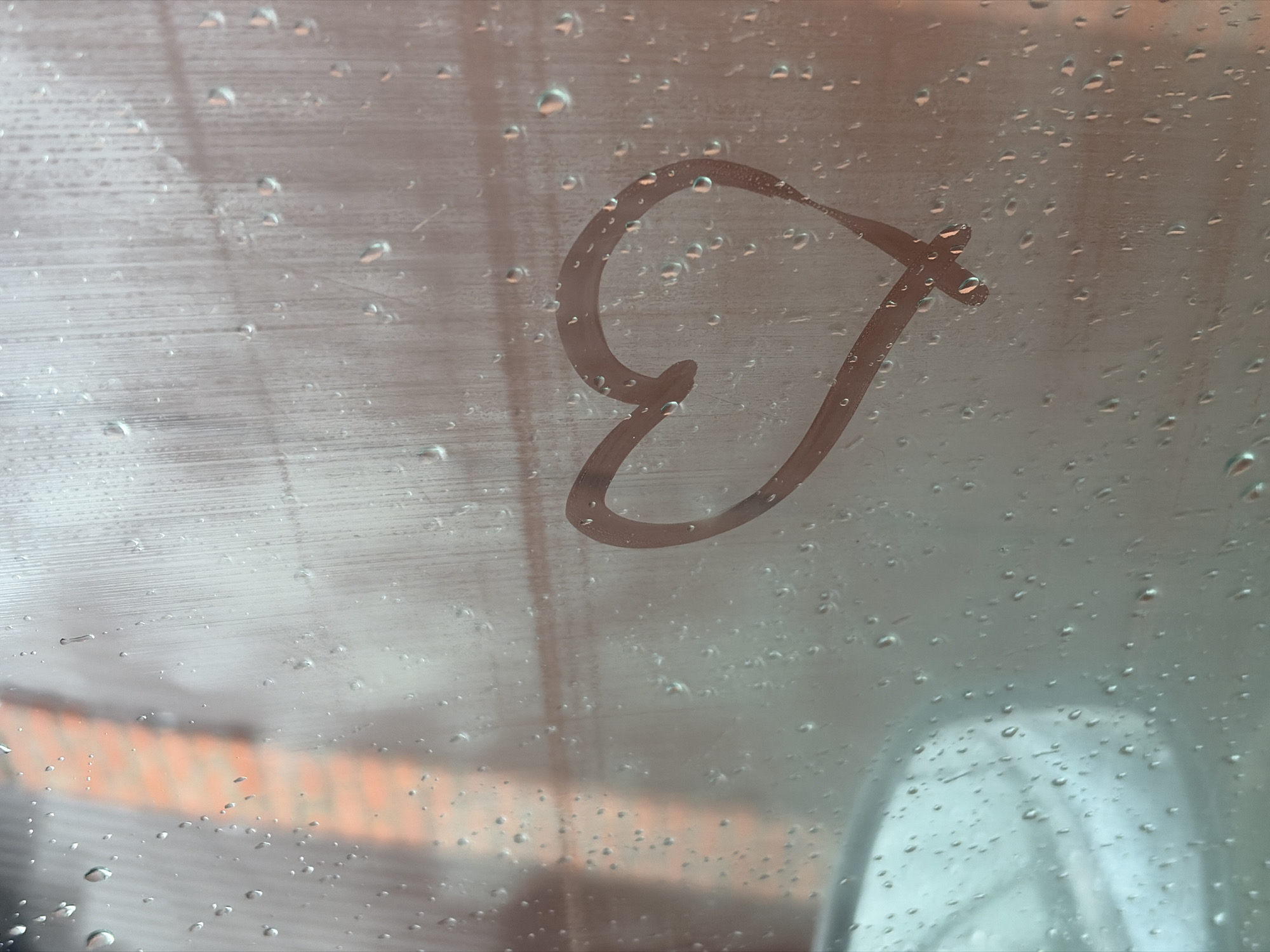 The image shows a heart shape drawn on a foggy or wet window. The window is covered in water droplets. Through the window, blurry shapes of objects and colors can be seen, possibly a building. The location is assumed to be Chaikovsky, Russia. There are no people visible in the image.