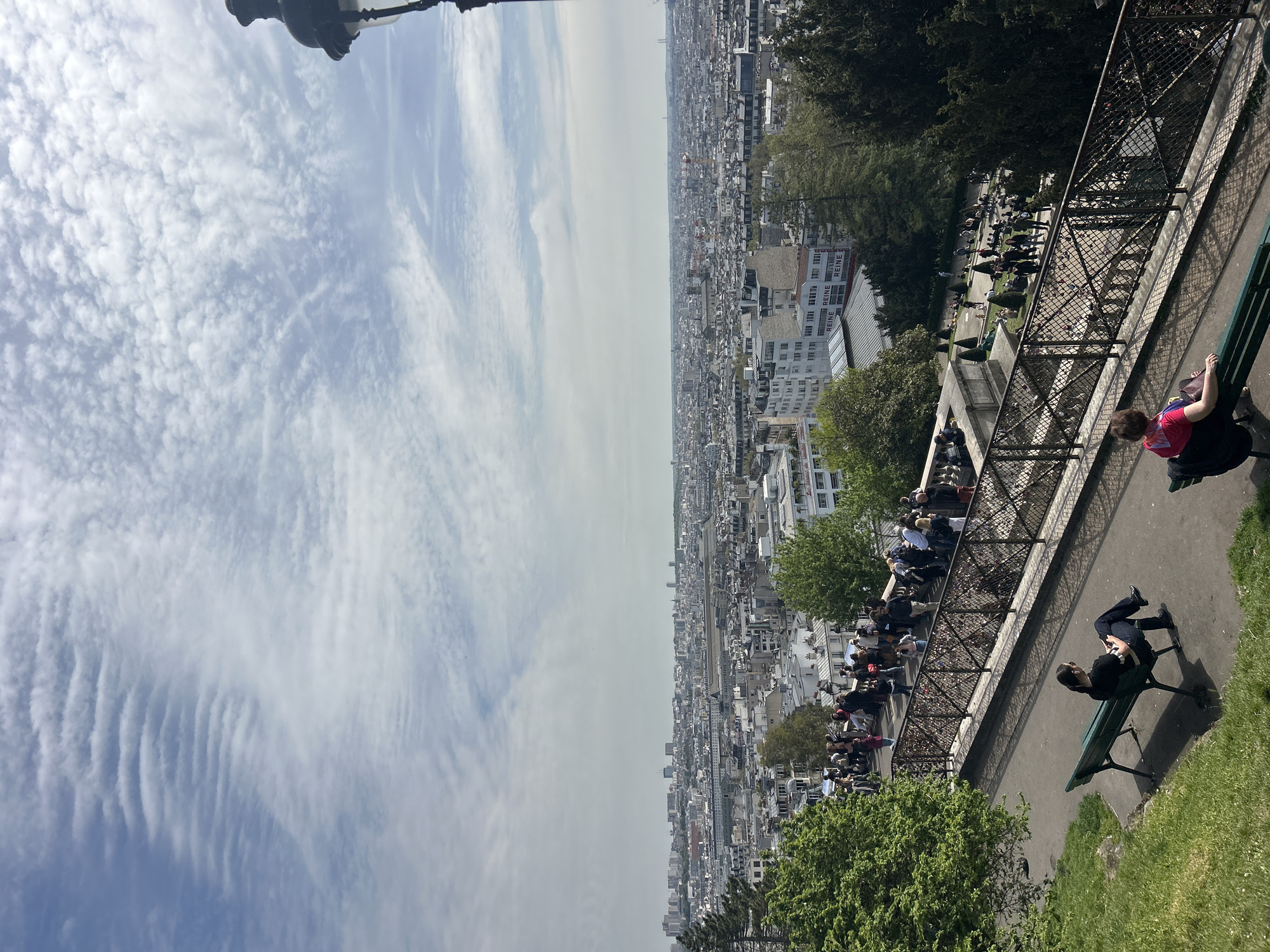 This image shows a cityscape view in Paris, France, likely taken from the area of Montmartre near the Sacré-Cœur Basilica. The frame includes a partial view of the sky with scattered cirrocumulus clouds. Below is a sprawling urban landscape with buildings of varying heights and architectural styles. Closer to the viewer is a park-like setting with trees, benches, and metal railings. People are visible, some seated on benches, others walking along a paved path. The image suggests a sunny day with tourists and locals enjoying the view of the city.