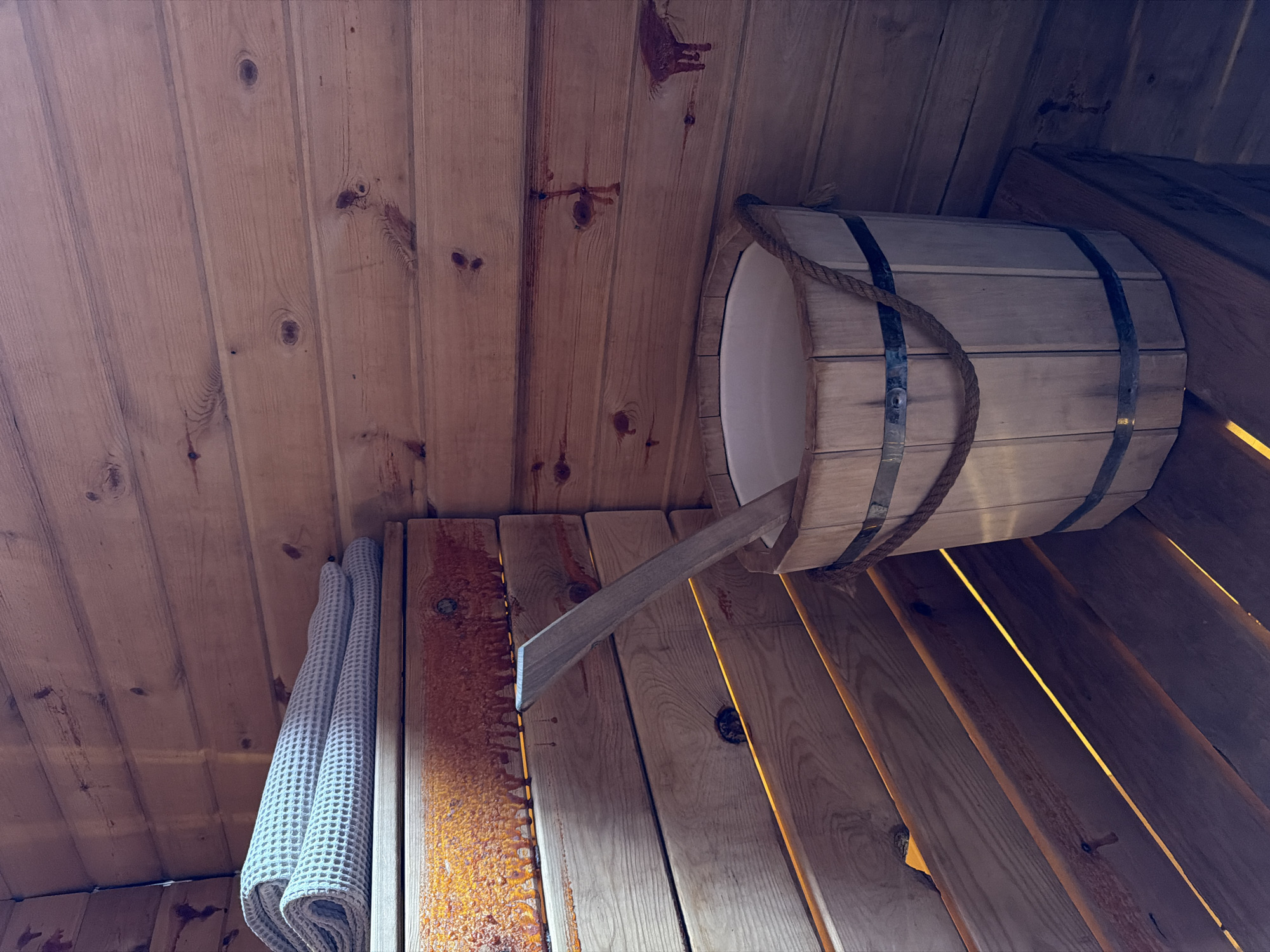 In an interior scene, a wooden bucket filled with water and a ladle sits on a wooden bench within a sauna. The walls and ceiling of the sauna are constructed from horizontal wooden planks. A neatly folded light-colored towel is placed on a higher wooden shelf. The lighting in the sauna is warm and inviting, with visible yellow light emanating from between the wooden slats. The overall environment suggests a relaxing and traditional sauna experience. The location of this scene is in the city of Семилуки, Russia.