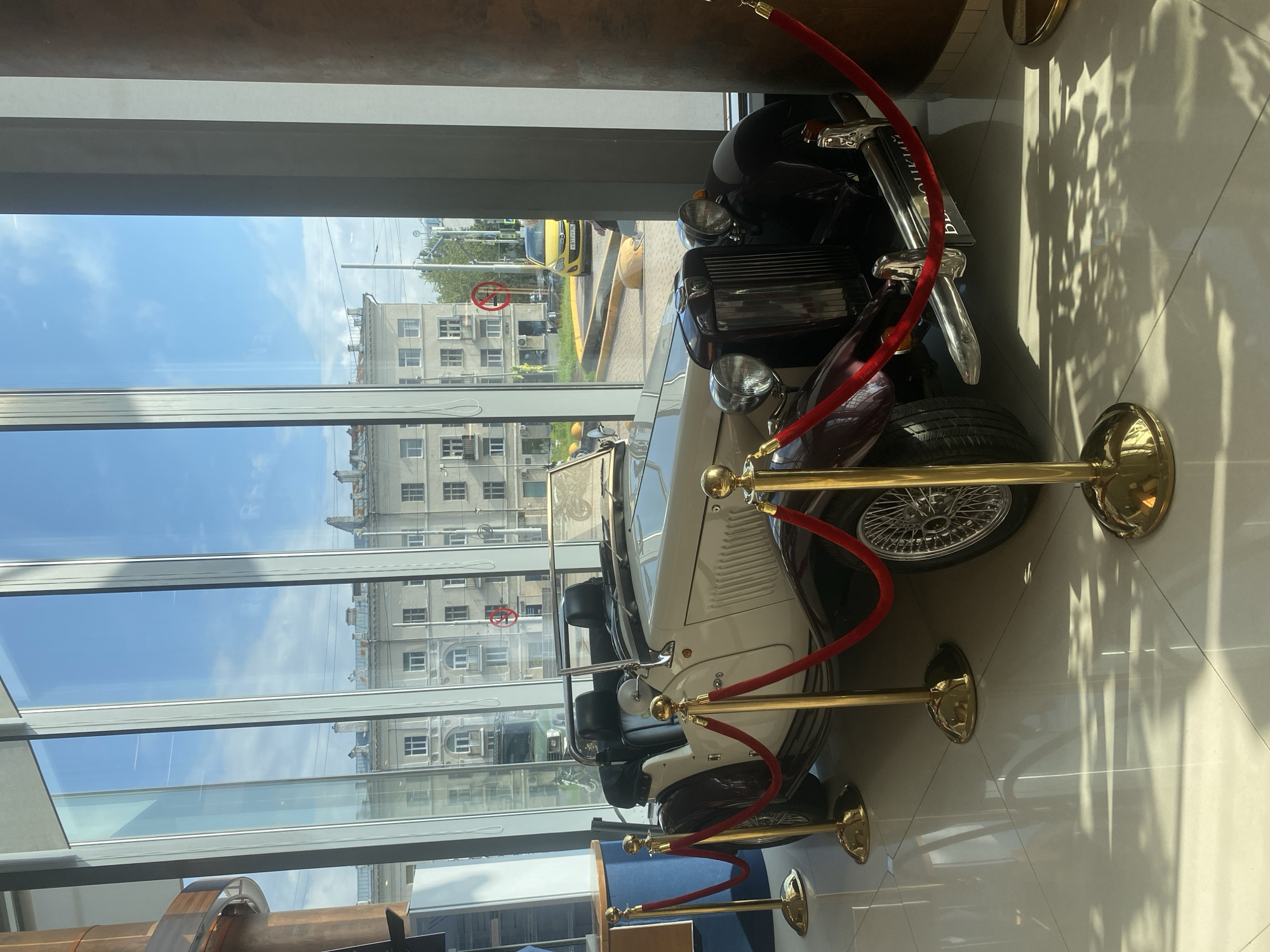 The image shows a white and dark red vintage convertible car parked indoors, with red velvet ropes and golden stanchions creating a barrier around it. The car is positioned in front of a large glass window that looks out onto a city street. Through the window, buildings, trees, a yellow car, and traffic lights are visible, suggesting an urban environment. The interior appears to be a modern building with a tiled floor that reflects the sunlight and shadows. The architectural style of the buildings outside, combined with the license plate of the yellow car which appears to have Cyrillic characters, suggests the location is likely in Russia, specifically the city of Yekaterinburg. The car itself is a classic roadster, likely a MG TD or similar model, with wire wheels and a convertible top. There are no people visible in the frame.