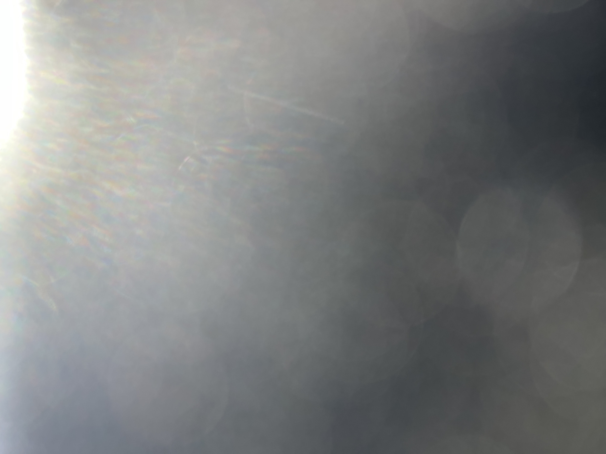 The image is an abstract, out-of-focus shot of light reflecting off a surface. There are no discernible objects, people, or environmental details. The dominant features are soft, circular blurs of light and faint streaks of color, suggesting a lens flare or bokeh effect. The overall tone is a gradient from bright white at the top to dark gray at the bottom.