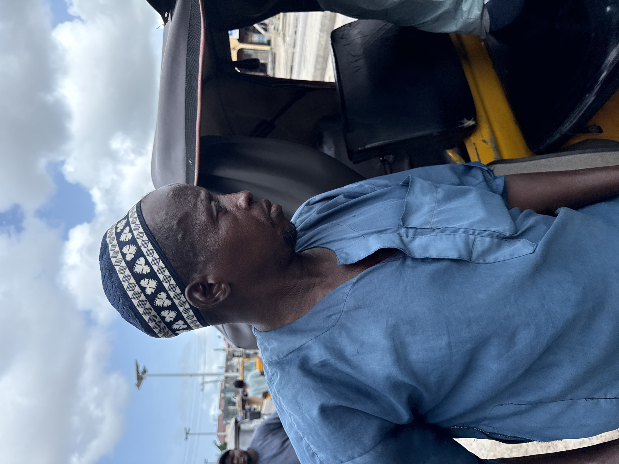 A medium shot of a man in a blue shirt and patterned cap in an auto rickshaw, viewed from the side. The man is likely seated inside the rickshaw. The background shows other rickshaws, utility poles, and a cloudy sky, suggesting an urban environment, possibly a vehicle park. The man has a beard and is wearing a blue, loose-fitting shirt. He appears to be looking ahead, though his gaze is slightly downward. The photograph gives a sense of transportation in a city and possibly a break time or waiting moment for the man. The photograph appears to be taken on location in Ado, Nigeria.