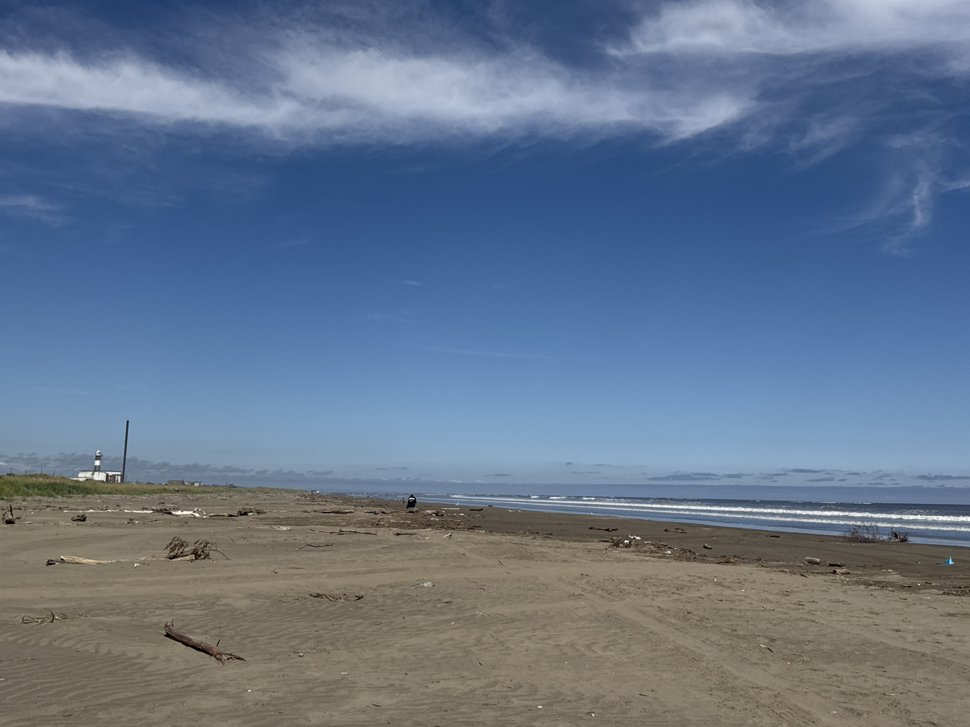 The image depicts a wide, sandy beach on a clear day. The sky is a vibrant blue with a wispy white cloud formation stretching horizontally across the upper portion of the frame. The sea is visible in the distance, with gentle waves rolling towards the shore. The beach is littered with driftwood and debris, indicating a tideline. In the far left of the image, a lighthouse with black and white markings stands tall. Further along the coastline, several small buildings can be seen. One person is visible on the beach, walking near the water's edge, dressed in dark clothing and a hood. The sand shows tire tracks, suggesting vehicle access to the beach. The overall scene is one of a natural, somewhat rugged coastline.