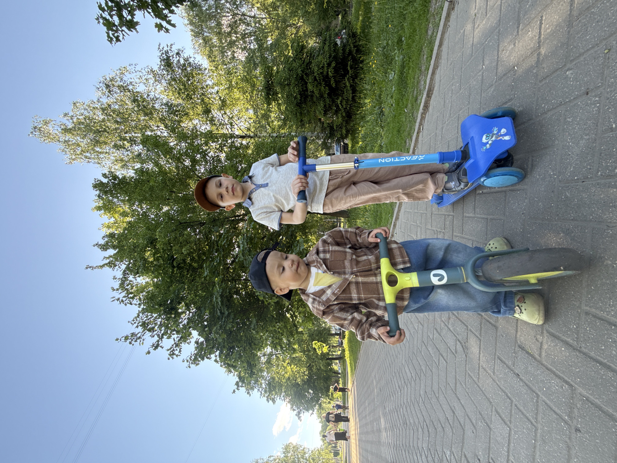 Two young boys are on a paved path in a park-like setting. The boy in the foreground wears a baseball cap, a brown plaid shirt over a yellow t-shirt, and jeans. He is riding a balance bike with green accents. The other boy is further away, wearing a brown cap, a beige polo shirt, and khaki pants. He is riding a blue scooter. Trees and grass are visible in the background. The sky is blue.
