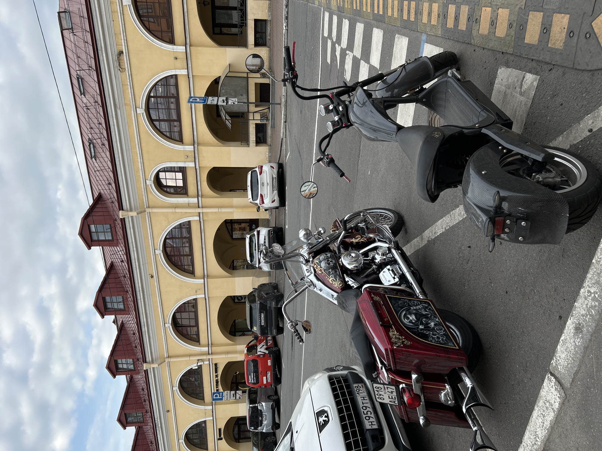 The image presents a street scene in Saint Petersburg, Russia, featuring a yellow building with a red roof and arched windows. In the foreground, there are two motorcycles. The first is a custom chopper in red and silver, adorned with skull-themed art, parked in front of a white car, its license plate visible and identifiable as "H959TH 198". The second motorcycle, located behind the first one, is a black scooter with a carbon fiber pattern. Several parked cars of various colors can be seen in the background, including a red van with "Яндекс Доставка" on it.
The architectural style of the building suggests a historical structure. A street sign with parking information is posted nearby. The road is marked with a yellow and white crosswalk. The sky is overcast.