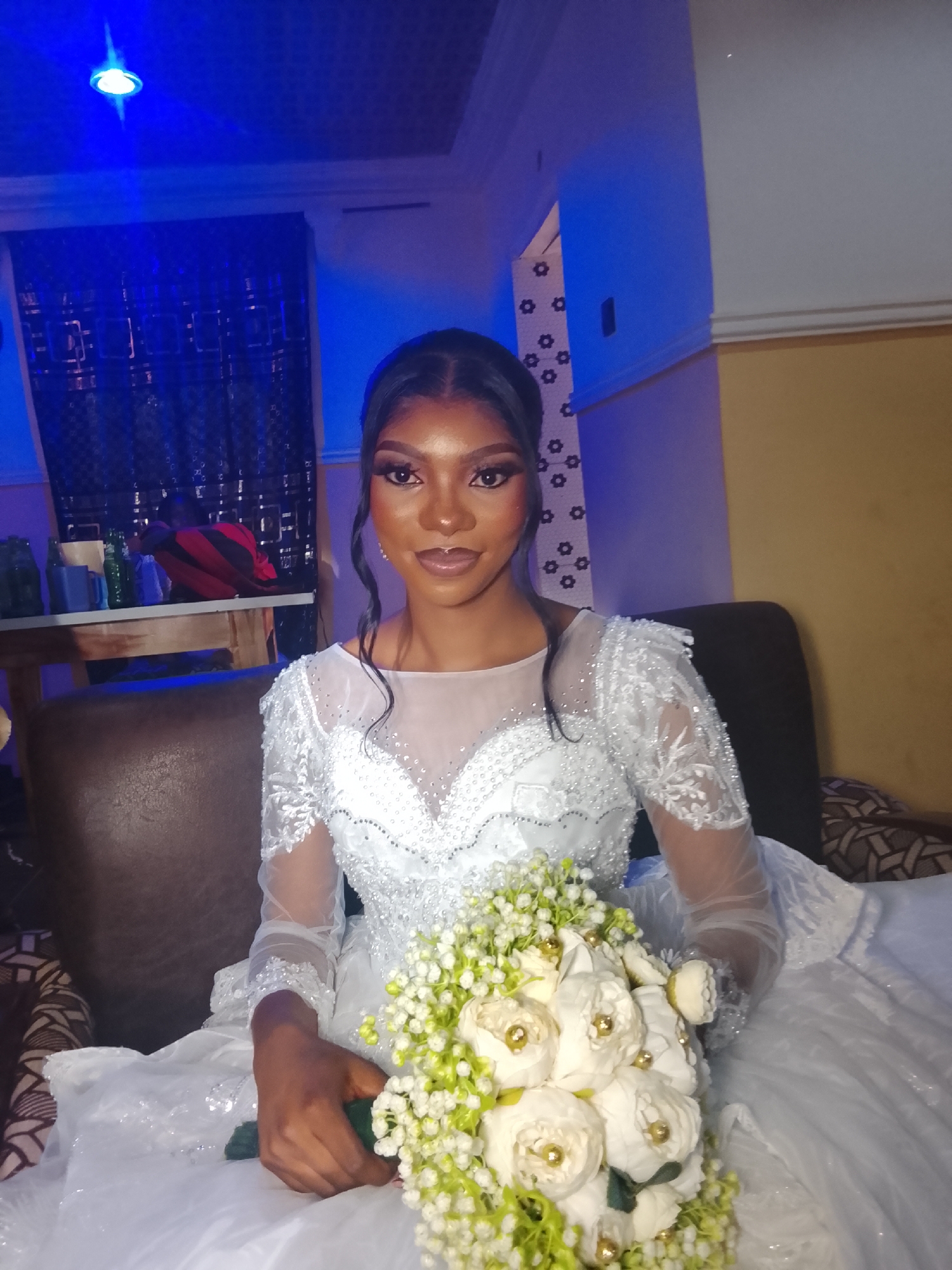 A woman sits on a couch holding a bouquet of white flowers. She is wearing a white, long-sleeved wedding dress. The location appears to be indoors in a residential area, likely Dogon Dinia, Nigeria. There is a table with various items on it in the background and blue lighting in the room. The couch is brown leather and there is wallpaper with a floral pattern on the wall.