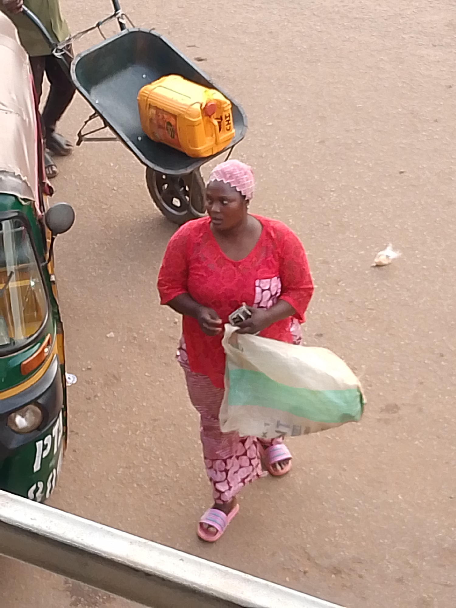 An overhead view captures a bustling street scene in Bukuru, Nigeria, featuring a woman, a man with a wheelbarrow, and an auto-rickshaw. In the foreground, a white barrier suggests the vantage point is from an elevated position looking down onto the road.
The central figure is a woman walking across the frame. She wears a vibrant red short-sleeved top, pink patterned trousers, and a matching pink headscarf. Her footwear consists of pink slide sandals. She carries a large, light-colored bag with a green stripe in her left hand and a small, dark object, likely a mobile phone, in her right.
To her left, a green and black auto-rickshaw (Keke Napep) is partially visible. White text on its front includes characters resembling "P1" or "P/1" and numbers like "24" or "28". To the woman's right and slightly behind her, a man in a green shirt and dark trousers is seen pushing a dark-colored wheelbarrow. Inside the wheelbarrow rests a bright yellow jerry can with a red cap, clearly marked with the letters "CHI" on its side.
The ground is a light brown, unpaved surface, with a small white plastic bag discarded near the woman. The scene depicts everyday activities under natural daylight, with no discernible strong shadows or specific weather conditions.