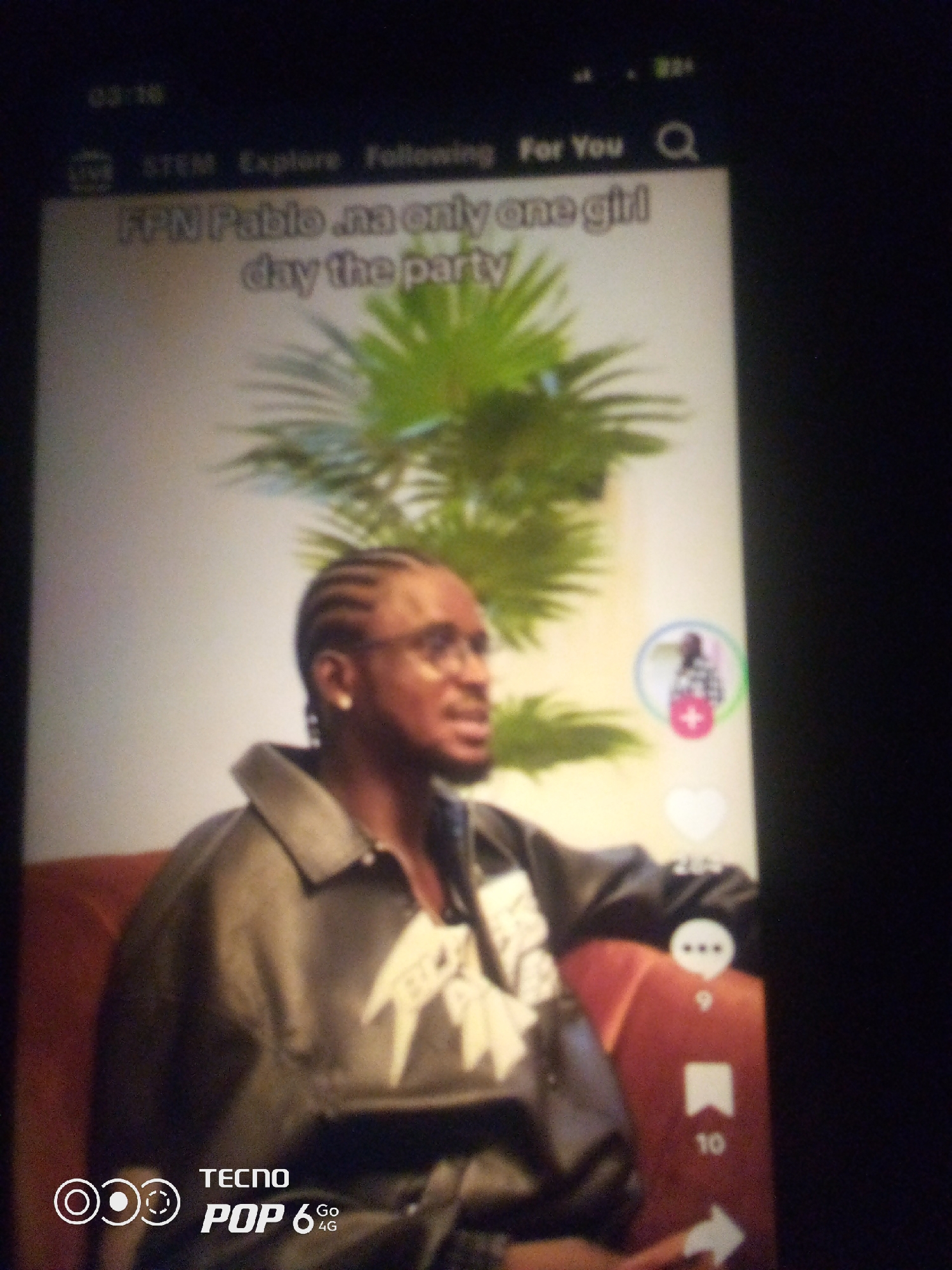 A man with braids and glasses is shown in a portrait. He is wearing a black jacket over a white shirt. The background features a large green plant and a light-colored wall. On the right side of the image, a profile picture with a plus sign is visible. The bottom left corner of the image displays the text "TECNO POP 6 4G". The man appears to be indoors, possibly in a studio or a residential setting. There is no indication of the city of Howayi, Nigeria.