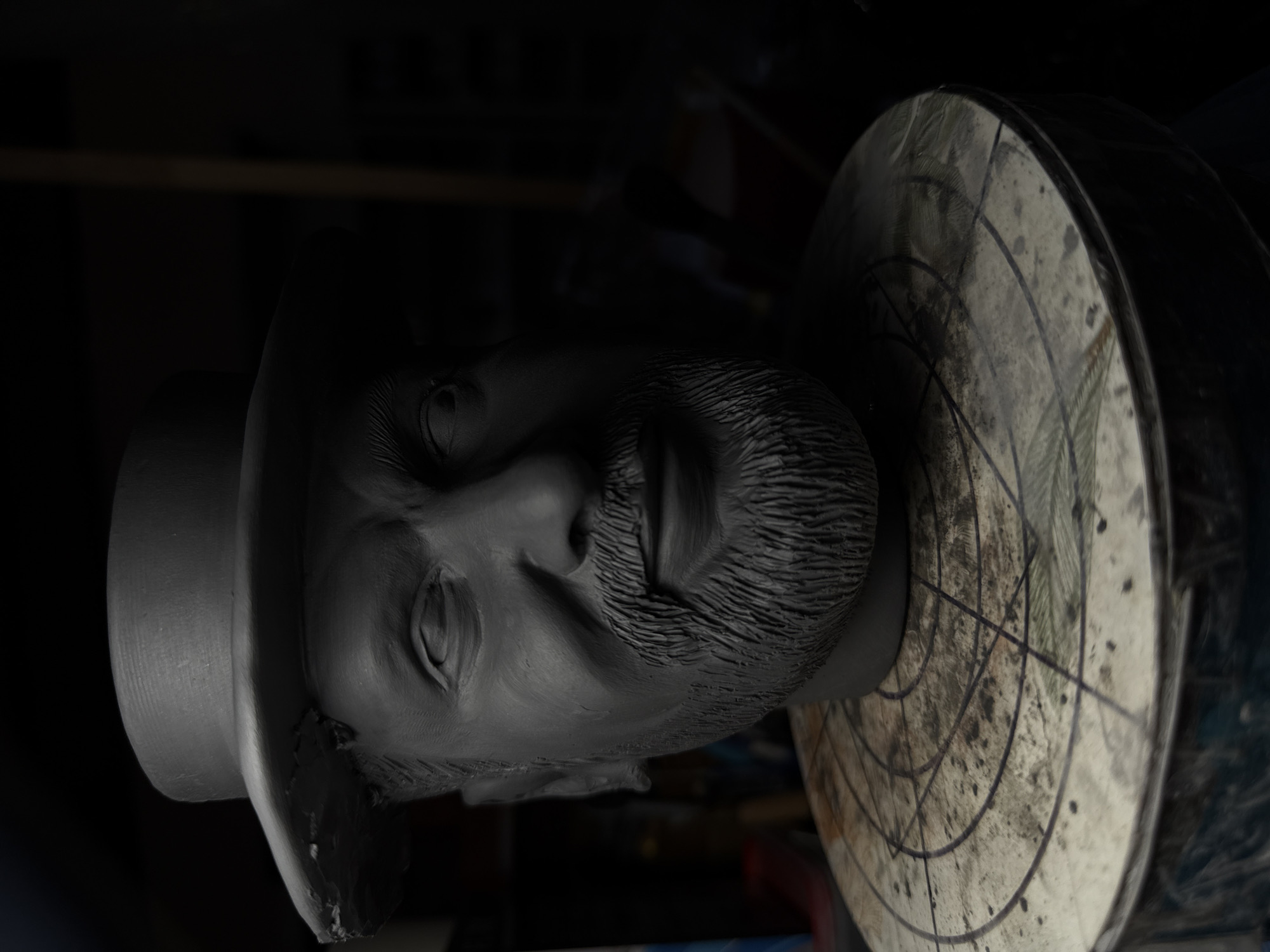 A grayscale, close-up shot shows a sculpture of a man's head wearing a hat, resting on a turntable. The head is sculpted with detailed facial features, including a beard and eyes. The hat is a simple design. The turntable is circular and has markings on its surface. The background is dark, with the subject appearing to be in a dimly lit studio. The sculpture is presumed to be located in the city of Chaikovsky, Russia.