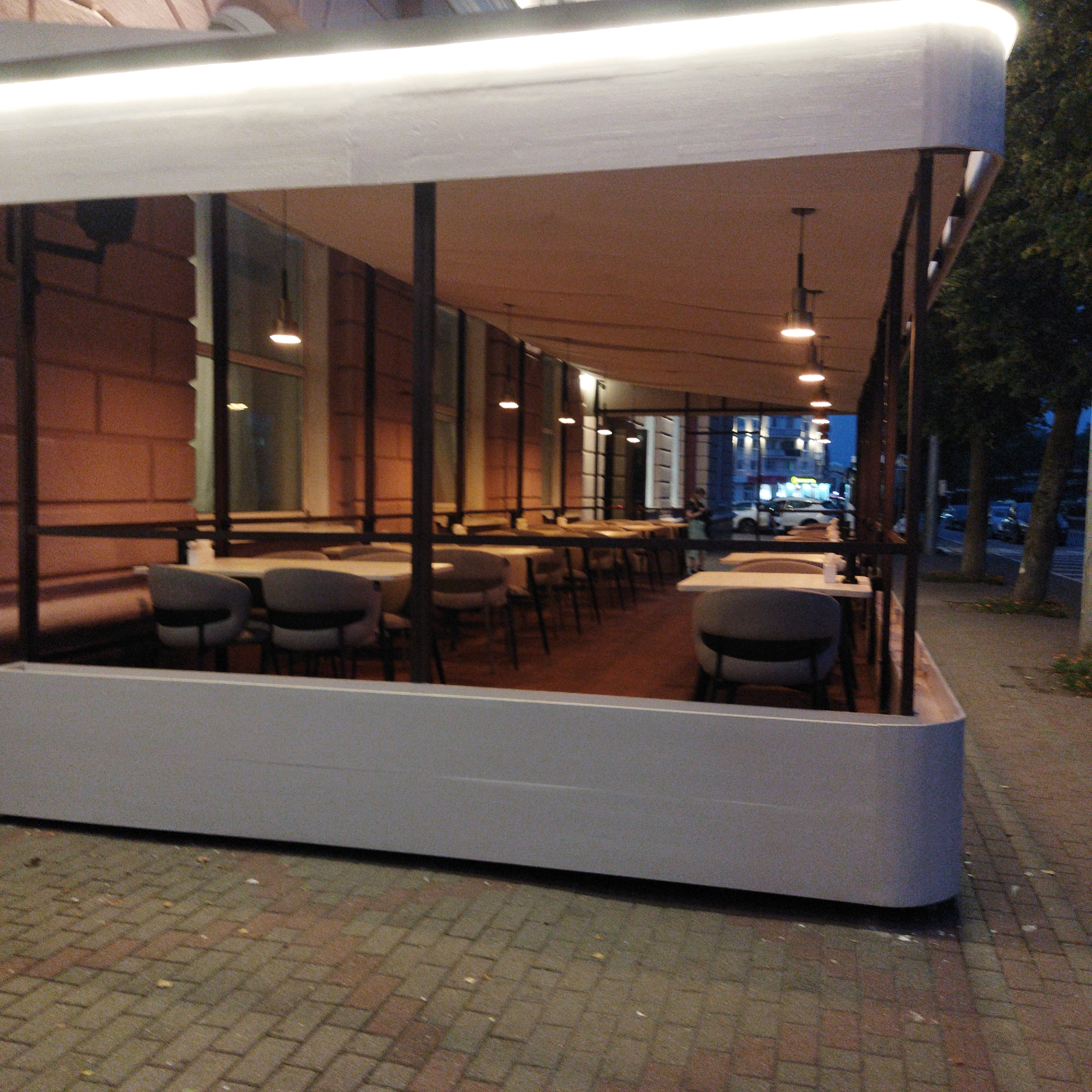 The image shows the exterior of a restaurant or cafe with an open-air seating area at dusk. The seating area is covered by a white canopy with integrated lighting. Several tables and chairs are arranged within the seating area. The facade of the building behind the seating area appears to be made of red brick. The sidewalk in front of the seating area is paved with brick pavers. In the background, through the glass of the building, streetlights and building lights are visible, suggesting an urban setting. There are trees and a street with cars in the distance. One person is visible standing inside the establishment. The scene is lit by artificial lights from the establishment and streetlights.