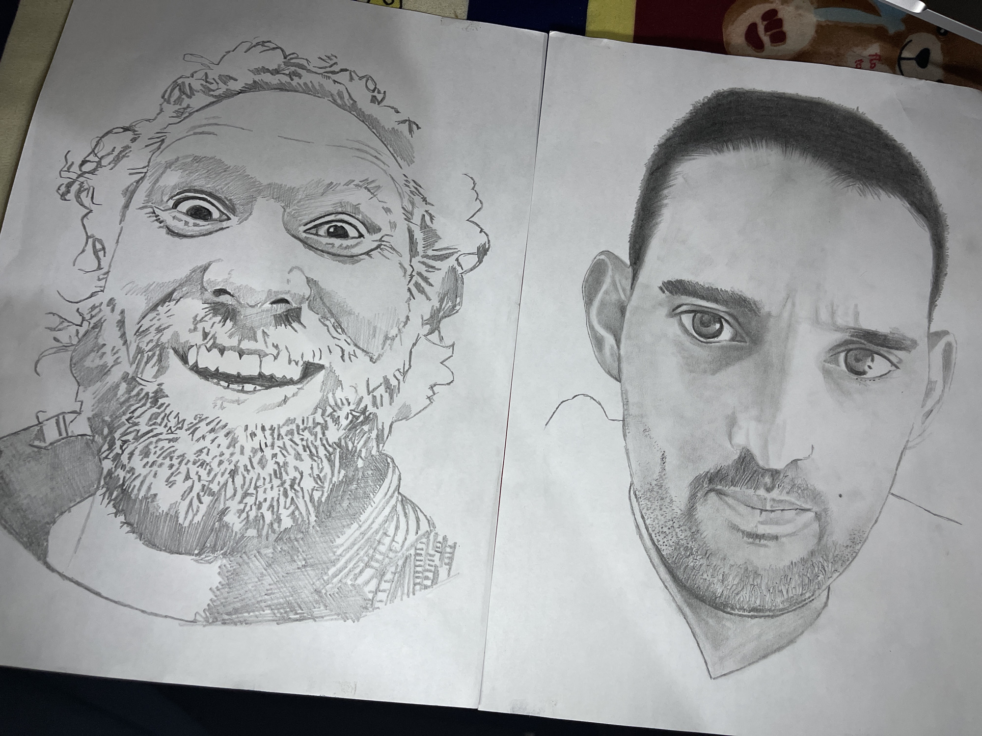 The image shows two pencil portraits on separate sheets of paper, likely in a notebook or sketchbook.
The portrait on the left depicts a man with a wide grin, visible teeth, a full beard, and curly hair. His eyes are wide and his expression is somewhat exaggerated. The shading is dense, particularly in the beard and hair, and uses cross-hatching to create texture.
The portrait on the right depicts a man with short hair, a neatly trimmed beard, and a more serious expression. His eyes are also emphasized. The shading is smoother and more even than in the left portrait.
The background is a blurred image which includes bedsheets, pillows, and other household items, suggesting the photo was taken indoors.
There is no evidence to suggest that this image was taken in Jos, Nigeria.