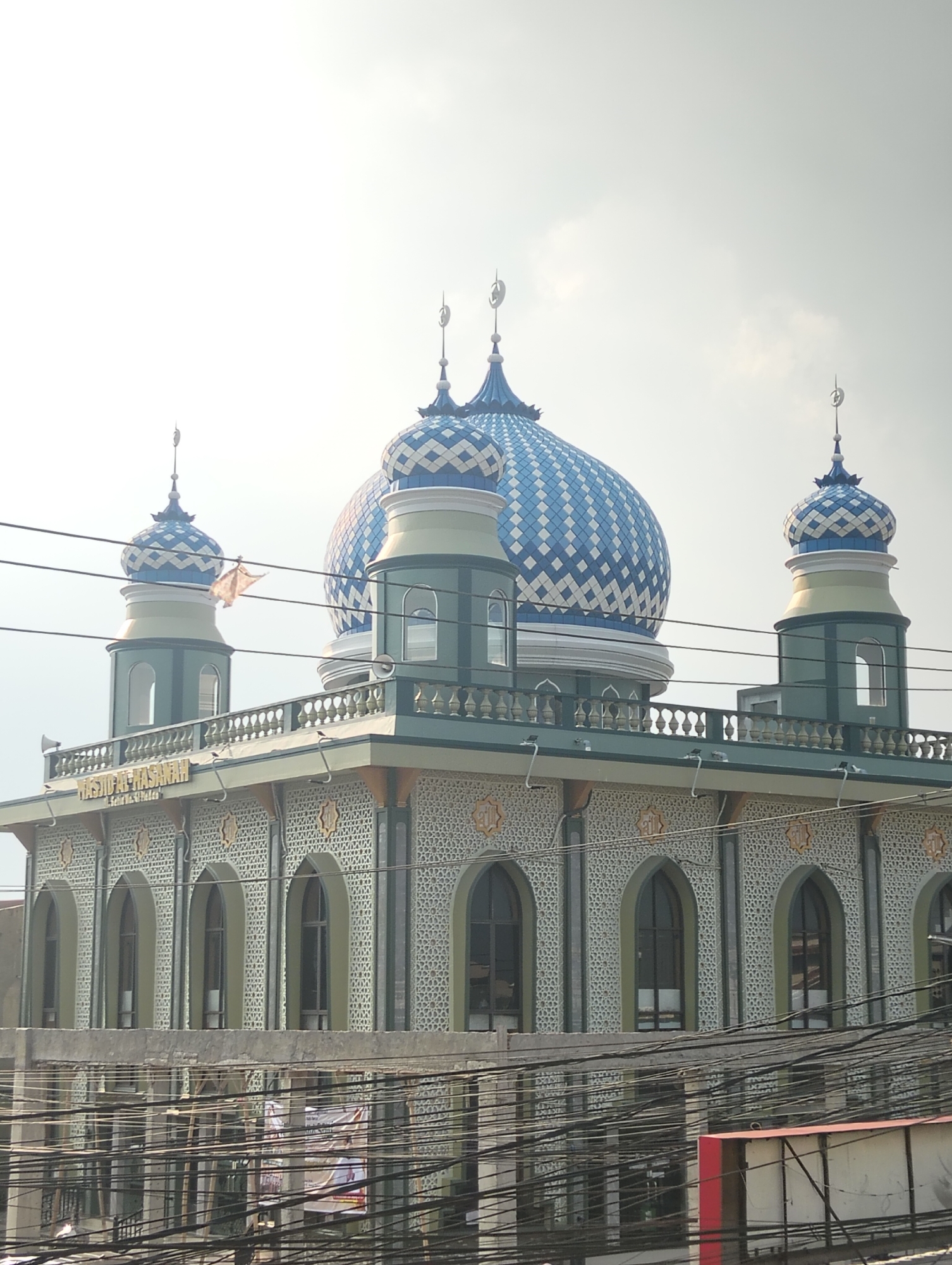 In the city of Medan, Indonesia, a mosque named Masjid Al-Hasanah is visible, characterized by its prominent blue and white checkered dome and two smaller matching minarets. The main structure of the mosque is green with ornate, patterned walls and arched windows. A balcony with decorative railings runs along the top of the building. Overhead, a tangle of black electrical wires obstructs the view of the sky and the upper portions of the mosque. The scene is captured from a slightly elevated perspective, with a bright, overcast sky in the background.