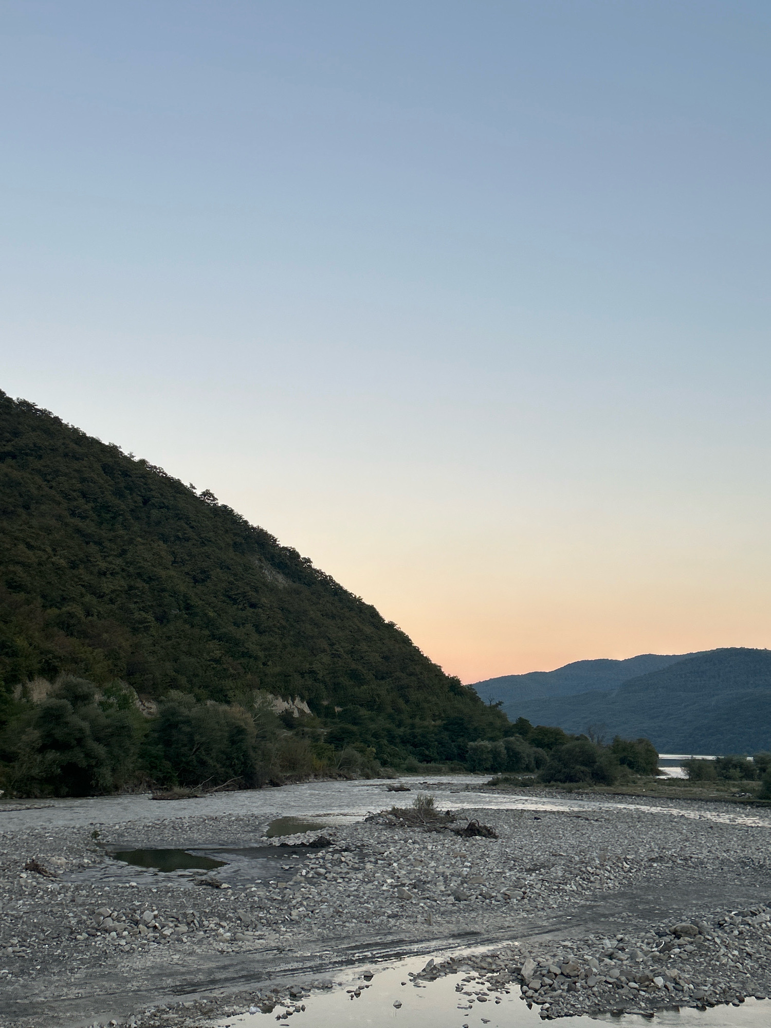 The image captures a serene natural landscape in Pavleuri, Georgia. A wide river, characterized by a stony bed and calm water, flows through the scene. On the left, a steep, tree-covered mountain slopes down towards the river. Across the river, in the middle ground, more trees and vegetation are visible, separating the river from a range of distant, undulating mountains. The sky above is clear, transitioning from a pale blue at the top to a soft orange hue near the horizon, suggesting it is either sunrise or sunset. There are no people or animals visible in the frame. The overall impression is one of natural tranquility.