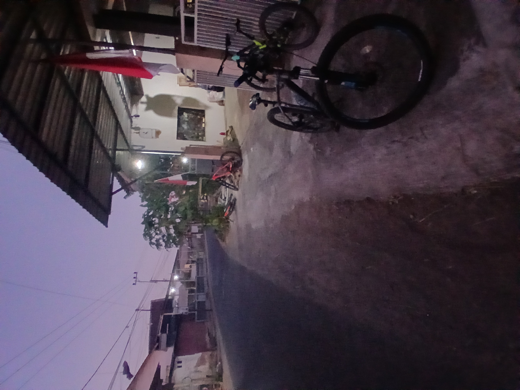 The image depicts a street scene in Kademangan Kulon, Indonesia, during twilight. Two bicycles are prominently parked on the concrete walkway in the foreground, one black and the other dark with yellow accents. A red and white Indonesian flag hangs from the corrugated metal roof of a building to the right. Behind the bicycles, there are more bicycles parked near the building. The street itself is paved and curves to the left, lined with modest buildings and power poles. The sky is a soft purple and pink hue, indicative of dusk.