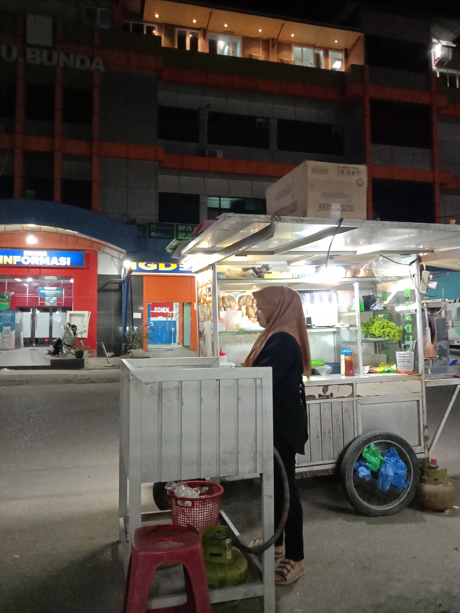 At night in Lhokseumawe, Indonesia, a woman wearing a hijab stands by a food cart on a street. The cart is white, with a box on top. Behind the cart, there are two buildings: a red and blue "Informasi" building, and an orange building. Two people are sitting on a bench near the "Informasi" building. A red stool and two propane tanks sit near the food cart.
