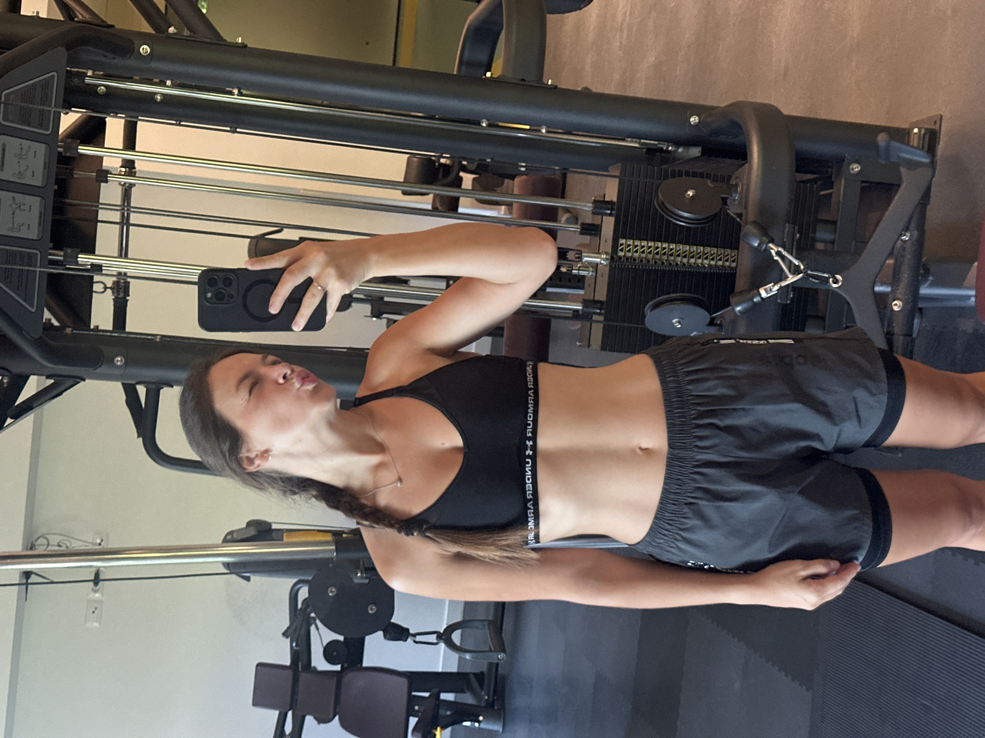 A young woman with her hair in a braid takes a selfie in a gym in Boreti, Montenegro. She is wearing a black sports bra and grey athletic shorts. In the background, a weight machine and other gym equipment are visible. The woman is puckering her lips and winking at the camera. The image is taken from the waist up.