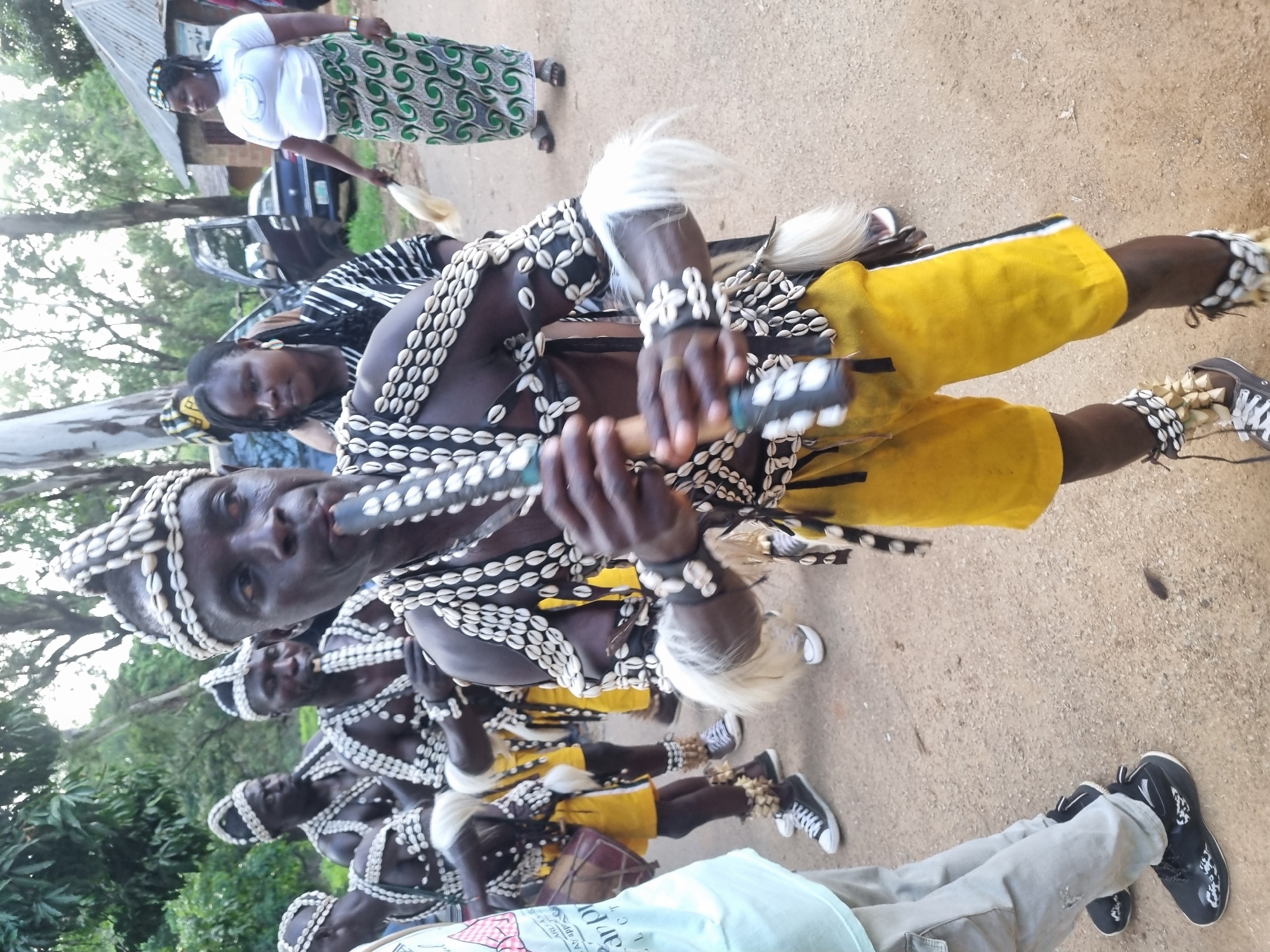 In Jos, Nigeria, a group of individuals are participating in a cultural ceremony. The foreground is dominated by a man dressed in traditional attire, playing a flute. His costume features a headdress adorned with shells, a necklace made of shells, and wristbands also decorated with shells and white fur. He wears bright yellow shorts and black and white sneakers. Behind him, other men in similar traditional outfits are visible, some also appearing to play instruments. To the right, two women are present. One woman, in the background, wears a white t-shirt with a logo and a green and white patterned skirt, holding a white object. The other woman, closer to the foreground, is dressed in a black and white striped garment and wears earrings. The scene is set outdoors on a dirt ground, with trees and a small building with a corrugated roof visible in the background. Cars are also present, suggesting a public gathering or event.
