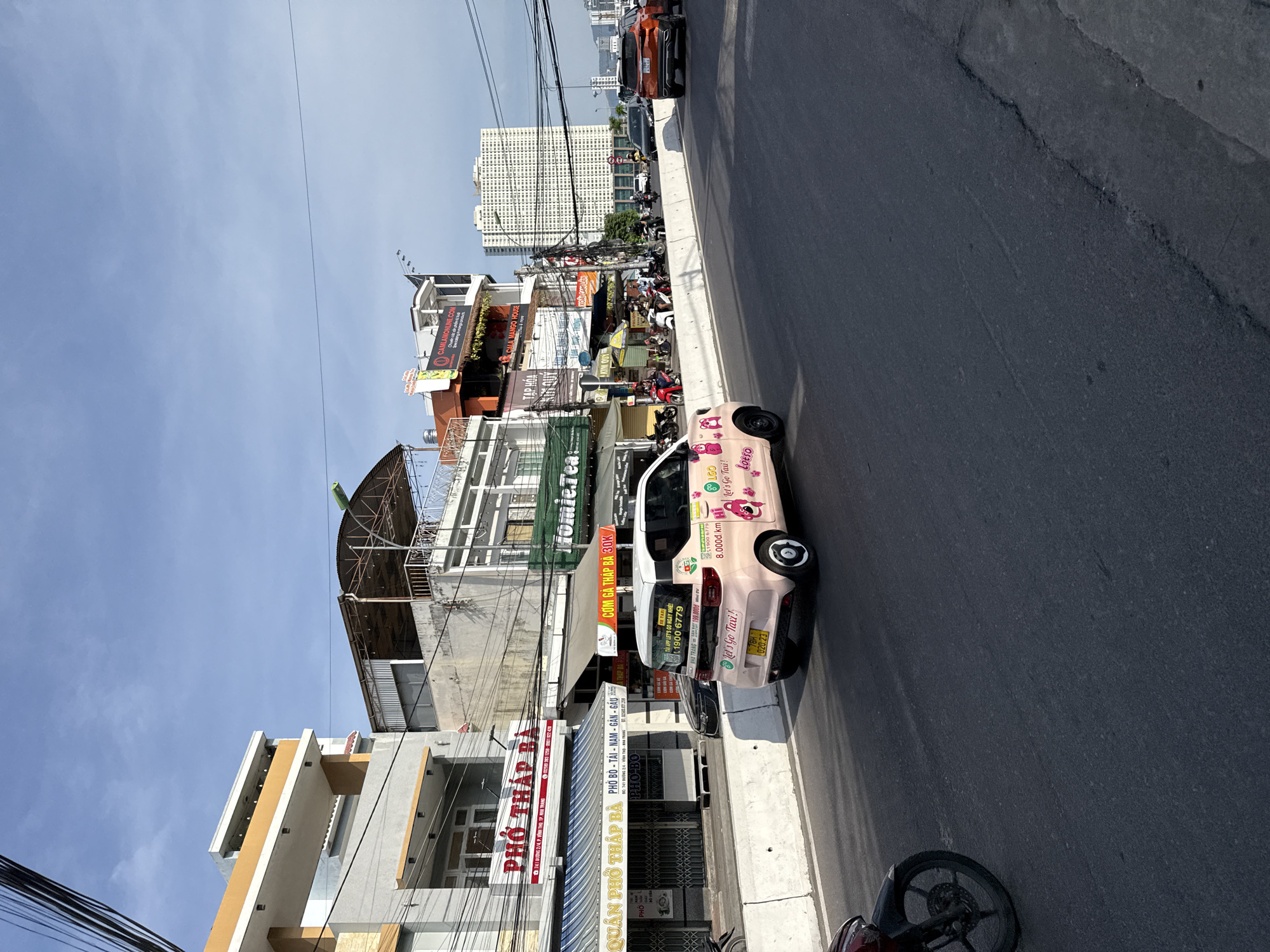 The image depicts a street scene in Bac Nha Trang Ward, Vietnam, under a clear blue sky with scattered clouds. A pink electric car, labeled "Lets Go Taxi" and featuring cartoon bear graphics, is driving on the road. The car has a license plate that reads "78H 020.23". In the background, several buildings line the street, displaying various signs in Vietnamese, including "PHO THAP BA" and "HOMIE.TEA". Other vehicles, such as motorcycles and cars, are visible on the road and sidewalk. Power lines crisscross the sky above the buildings. The environment suggests a bustling urban area with commercial establishments and ongoing traffic.