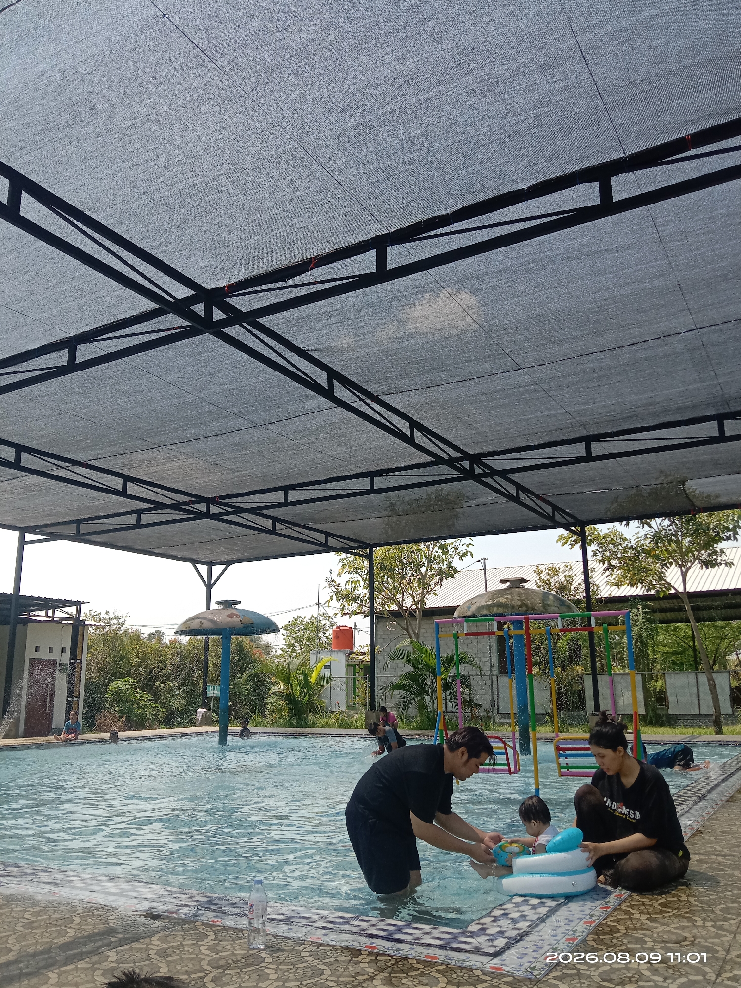 The image depicts a scene at a swimming pool, likely in Blimbing, Indonesia. In the foreground, a man and a woman are interacting with two young children in the shallow end of the pool. The man, wearing a black t-shirt, is holding a blue and white inflatable toy, seemingly assisting one of the children. The woman, also in a black t-shirt, is sitting on the pool's edge, looking towards the children. One child is wearing a blue inflatable ring in the water, and another child is partially visible.
The swimming pool has clear, rippling water and a tiled edge. A water dispenser with a mushroom-shaped top is visible in the pool, and a colorful playground structure with swings is present in the background. Several other people are in the pool and around its perimeter, some swimming or playing, while others are seated.
The area is partially covered by a gray mesh shade structure supported by black metal beams, suggesting an outdoor recreational facility. Lush green trees and buildings with white roofs are visible beyond the pool, under a bright sky. A water bottle is placed on the pool deck in the foreground. The timestamp "2026.08.09 11:01" is overlaid on the bottom right of the image.