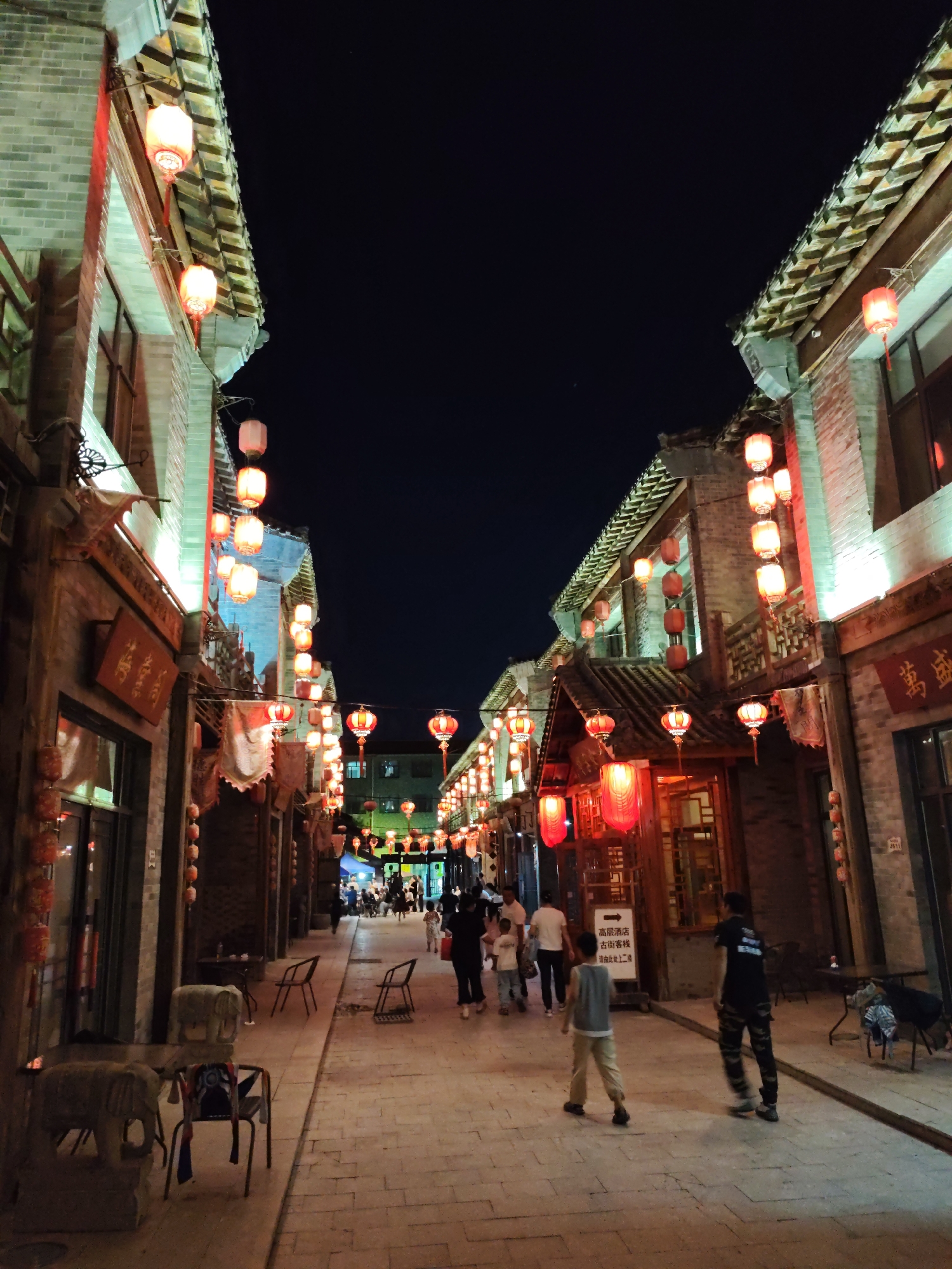 The image depicts a narrow street in Lindian, China, lined with traditional Chinese architecture, illuminated by numerous red lanterns at night. The buildings on either side of the street have tiled roofs and brick facades. People are walking along the brick-paved street, some in small groups, others individually. In the foreground, two children are walking away from the camera, and further down the street, more people can be seen, suggesting a lively evening scene. There are also chairs and decorative elements, such as elephant sculptures, placed along the sides of the street, possibly for seating or as part of the street's ambiance. The overall impression is one of a cultural and festive atmosphere.