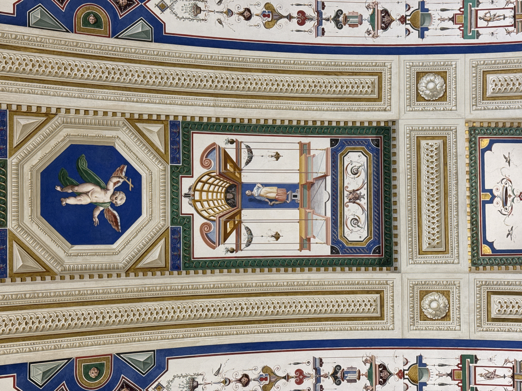 The image displays a highly detailed, ornate painted ceiling, likely an architectural fresco, found in San Lorenzo de El Escorial, Spain. The ceiling is divided into numerous geometric panels and borders, featuring a rich color palette of blues, golds, whites, reds, and greens.
Dominating the upper central octagonal panel is a fresco of a winged cherub, depicted in an upside-down pose, with green drapery trailing behind it. Other panels showcase intricate decorative motifs, including geometric patterns, stylized floral elements, and trompe-l'œil architectural details. Several rectangular panels contain depictions of mythological or allegorical figures, often rendered in a classical style, standing within architectural niches or elaborate frames. These figures include a woman in a yellow garment and a male figure in a warrior-like stance. Smaller details include representations of birds, vases, and what appear to be decorative lamps or urns. The overall impression is one of opulence and refined artistic execution, consistent with Renaissance or Baroque decorative schemes found in palatial or religious structures.