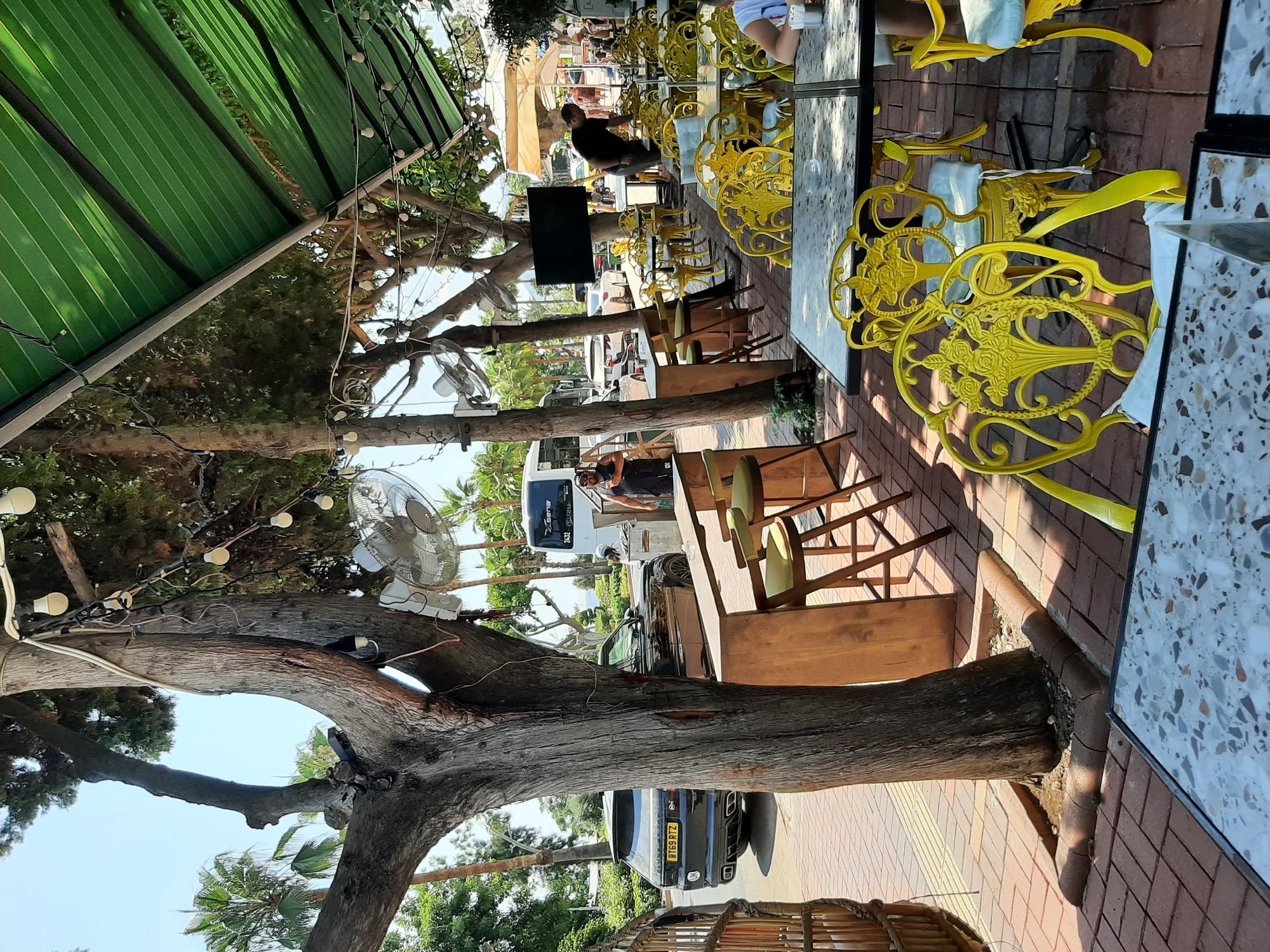 The image depicts an outdoor seating area of a cafe or restaurant, likely located in Alanya, Turkey, given the presence of palm trees and the general ambiance. A large, mature tree with a textured bark stands prominently in the foreground, its branches adorned with string lights and a small electric fan. Adjacent to the tree, there is a wooden counter with several green bar stools.
Beyond this area, rows of tables with decorative yellow chairs are arranged on a brick-paved patio. Some tables are covered with white cloths, and others have clear glass tops. A black Range Rover with a UK license plate (BT69 RTZ) is parked on the street to the left, with a pedestrian pathway separating it from the outdoor seating.
In the background, more tables and chairs are visible, along with umbrellas and awnings providing shade. Several vehicles, including a white bus or van with "ISUZU" branding, are on the street. Two men are present in the scene: one stands near the bus, looking at his phone, while another, wearing a black t-shirt and shorts, is observed attending to the tables. The scene is bathed in bright sunlight, suggesting a warm, daytime setting. The overall impression is a relaxed, outdoor dining or cafe environment, possibly during tourist season.