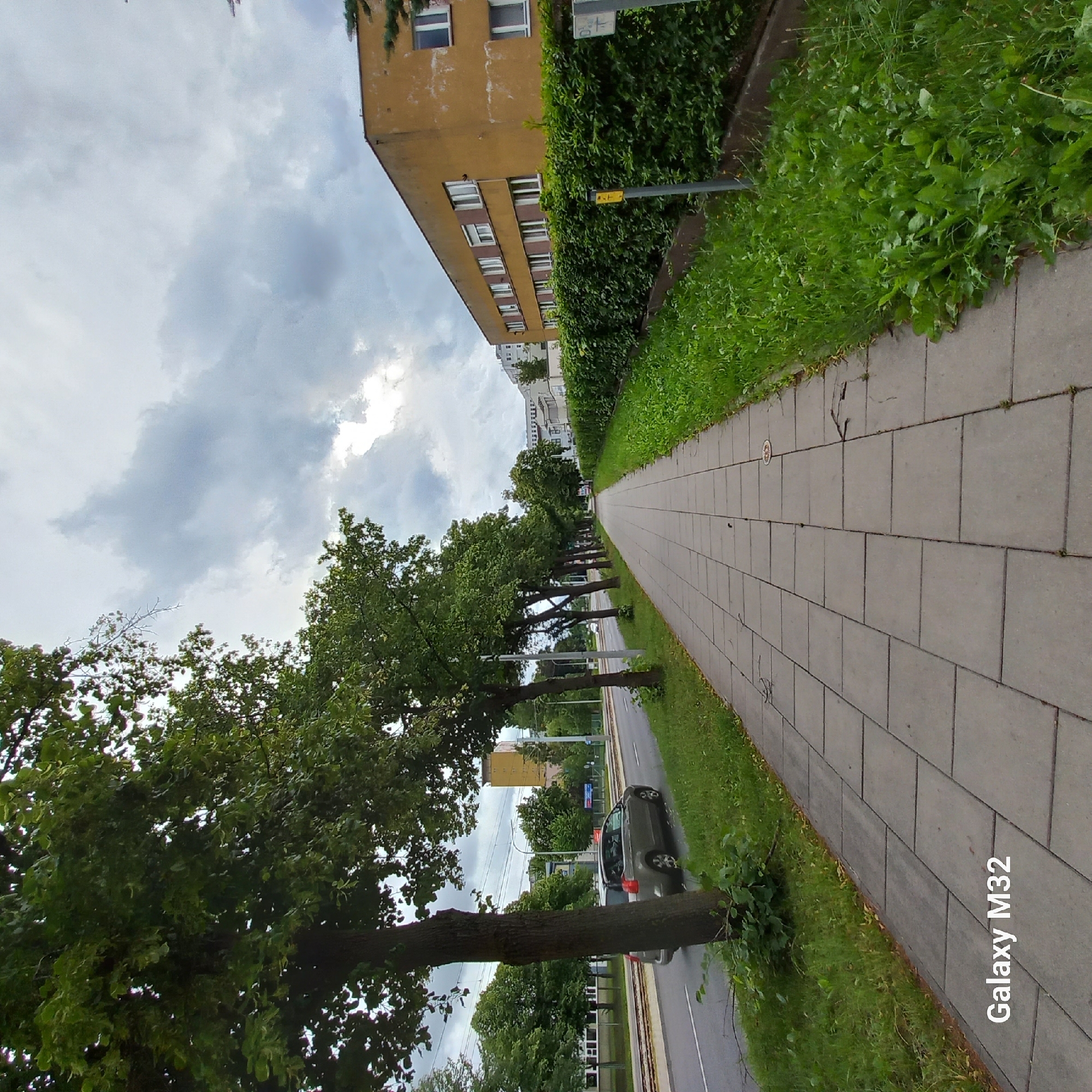 This is a full-frame, eye-level, outdoor, wide-angle photograph taken on a cloudy day in Olsztyn, Poland, likely during daytime. The scene depicts a paved pedestrian walkway lined with trees on the left and a dense green hedge with a yellow building behind it on the right. A street with tram tracks and a car is visible to the left of the walkway. The walkway is made of grey square paving stones, some of which show wear and tear. The grass verge to the right of the walkway is overgrown with various plants. The sky is filled with heavy grey clouds, with some lighter patches suggesting diffused sunlight. The yellow building on the right appears to be an apartment or office building with multiple windows. The overall atmosphere is that of a residential or urban street with public amenities. The bottom left corner of the image displays a watermark that reads "Galaxy M32".