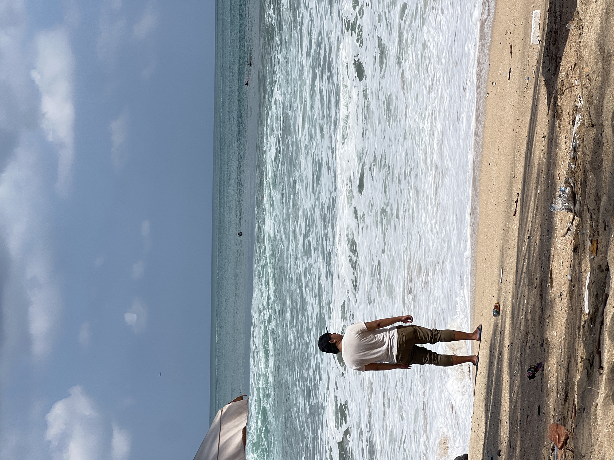 A person stands on a sandy beach in Kuta, Indonesia, facing the ocean. The person is wearing a light-colored t-shirt and dark pants, with a dark baseball cap. The ocean is a turquoise green with white waves breaking on the shore. In the distance, several surfers are visible in the water. The sky is partly cloudy, with a few white clouds against a blue sky. The beach is littered with various debris, including plastic bottles and other trash, indicating a lack of cleanliness. The sun is casting shadows on the sand, suggesting it is daytime. The overall scene depicts a natural environment impacted by human presence and waste.