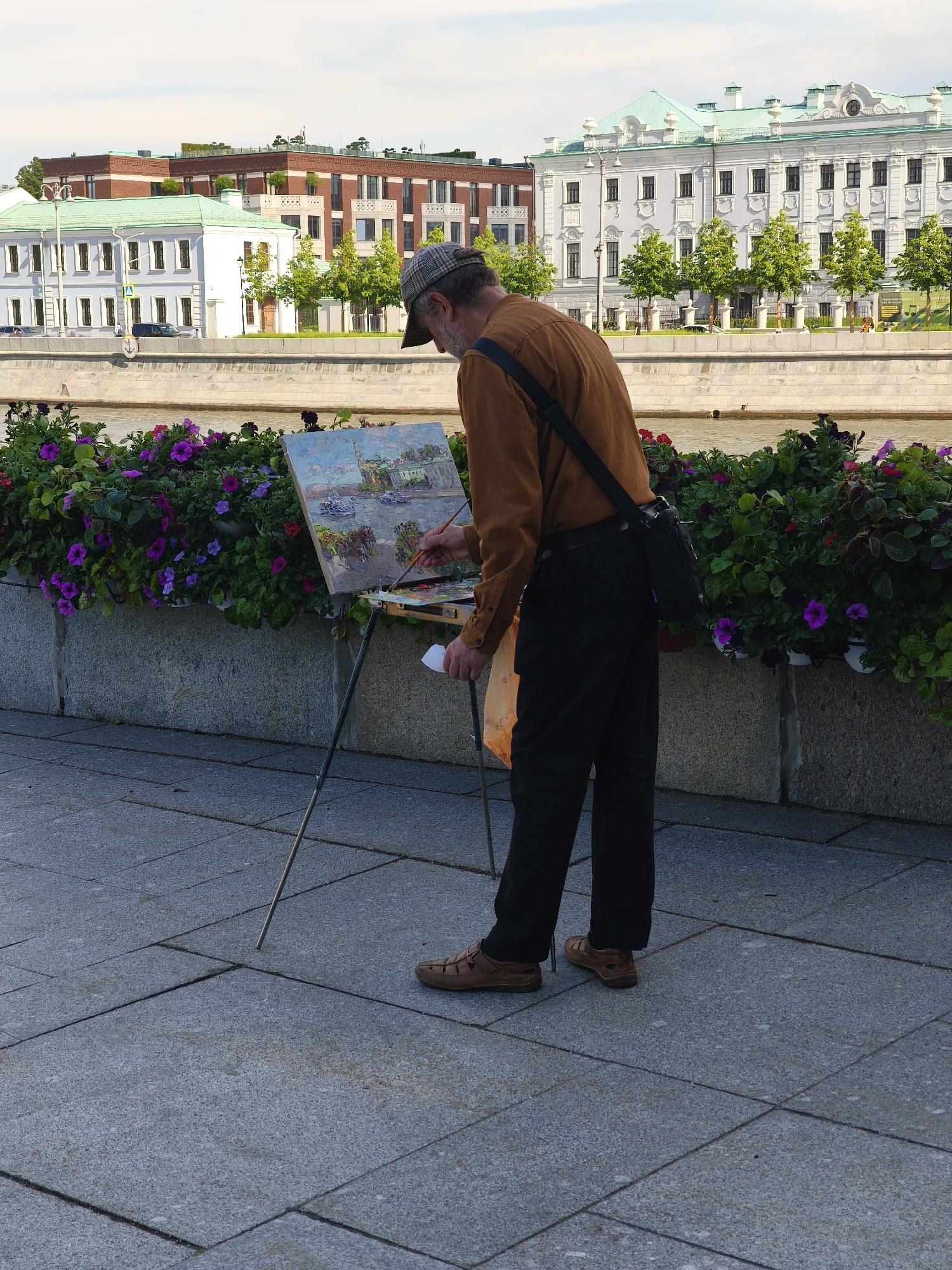 An outdoor, eye-level shot captures a male artist painting en plein air by a river on a sunny day. The artist, seen from the back and side, wears a brown long-sleeved shirt, black trousers, a plaid cap, and light brown shoes. A dark shoulder bag hangs across his body, and he holds a white cup and an orange plastic bag in his left hand while actively painting with a brush in his right hand. His canvas, mounted on a metal easel, depicts a landscape, likely of the scene before him, featuring buildings and water.
In the foreground, a paved stone sidewalk transitions to a stone embankment topped with planters overflowing with vibrant purple and pink petunias. Beyond the flowers, a calm river reflects the bright sky. On the opposite bank, several buildings are visible: a prominent white multi-story building with a light green roof and ornate detailing, and a larger brown building with red brick facades and multiple balconies, also with a green roof. Lush green trees line the riverbank between the buildings and the water. A small yellow pedestrian crossing sign (white figure on black stripes) is visible on the far bank, and a white anchor symbol is embossed on the embankment wall. The overall atmosphere is peaceful, capturing an artist immersed in his craft amidst an urban waterside setting.