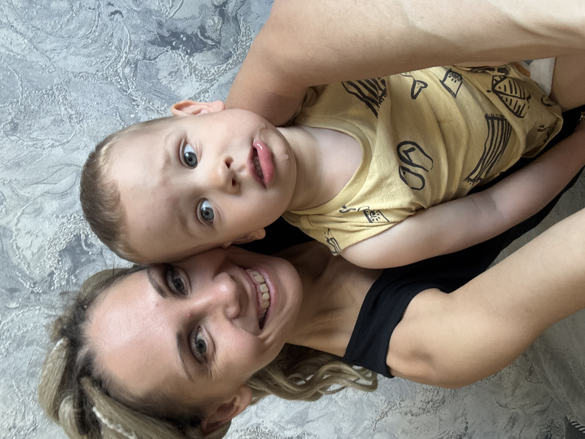 A selfie-style, slightly angled overhead shot captures a woman and a young child. The woman is lying on her back, smiling, with her face close to the camera. She has fair skin, light-colored hair pulled back with visible sections of braids, and is wearing a black top. The child, with fair skin and light hair, is positioned above her, facing the camera with an open mouth. The child wears a mustard-yellow shirt with dark graphic prints. The background is a textured gray and white surface, suggestive of bedding or a floor covering.
Given the prompt mentioning the city of Rusanovo, Russia, it can be inferred that the location of the image is potentially within a domestic interior in Rusanovo, but there are no specific landmarks or features visible in the image to confirm this.