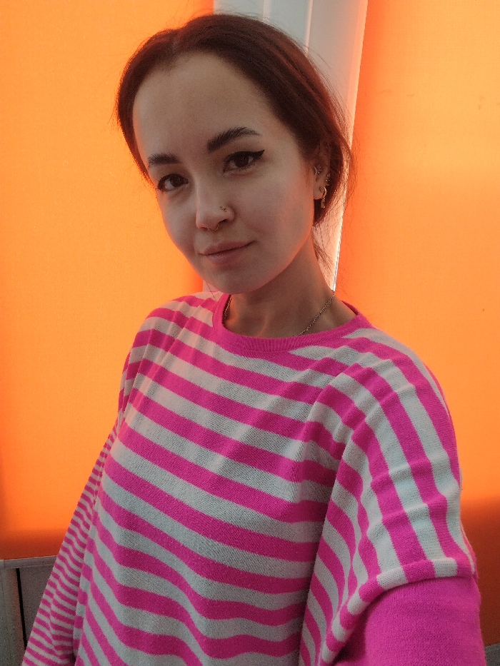 A young woman poses indoors with an orange background. She has dark hair, eyeliner, and a nose piercing. The woman wears a pink and white striped sweater with long sleeves. A silver chain is visible around her neck. Earrings adorn her left ear. She looks directly at the camera with a slight smile. The scene appears to be lit with bright, even lighting.