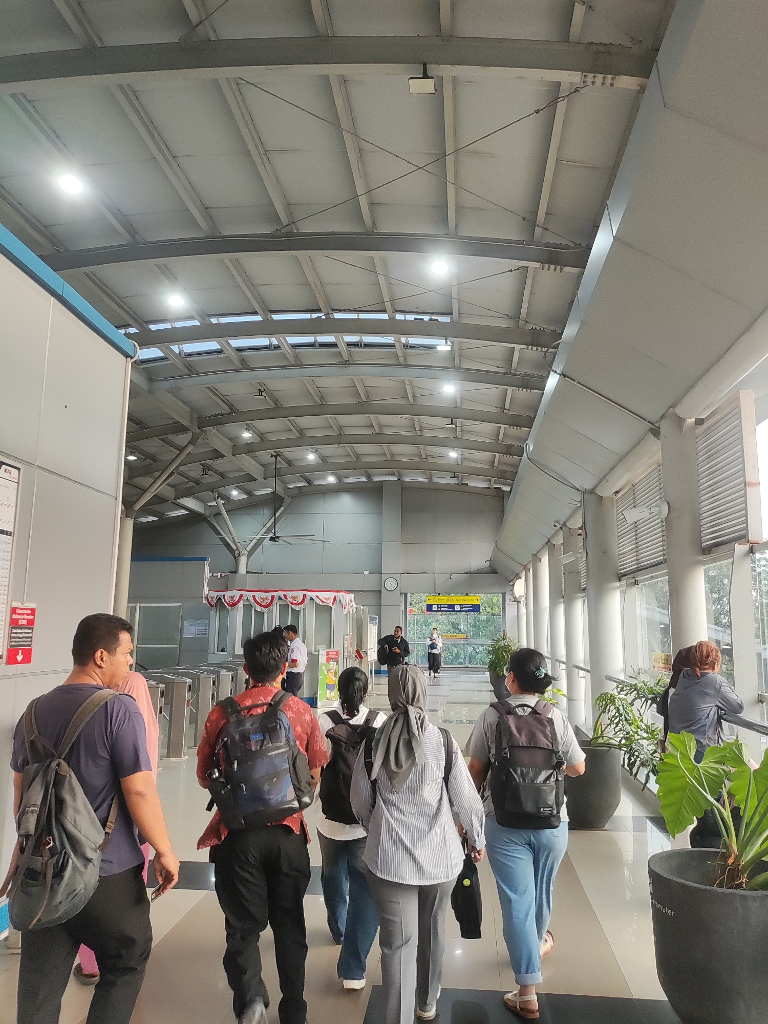 The image depicts an interior view of a modern transit station in East Jakarta, Indonesia, likely a train or bus station, characterized by a high, exposed ceiling with metal beams and translucent panels allowing natural light. The scene is bustling with people, predominantly dressed in casual attire, walking in various directions. Many are carrying backpacks, suggesting they are commuters or travelers.
In the foreground, a man with a backpack walks away from the camera, his head turned slightly to the left. To his right, another man in a red patterned shirt, also wearing a backpack, walks in the same direction. Behind them, a group of individuals are seen walking, including a woman in a striped shirt and headscarf, and another woman in a grey t-shirt and blue jeans, both with backpacks. Further ahead, the station features ticket gates and information displays. In the background, several people are visible near the gates and a small kiosk adorned with Indonesian flags. Large potted plants are strategically placed along the walkway, adding a touch of greenery to the urban environment. The overall atmosphere suggests a busy public transportation hub.