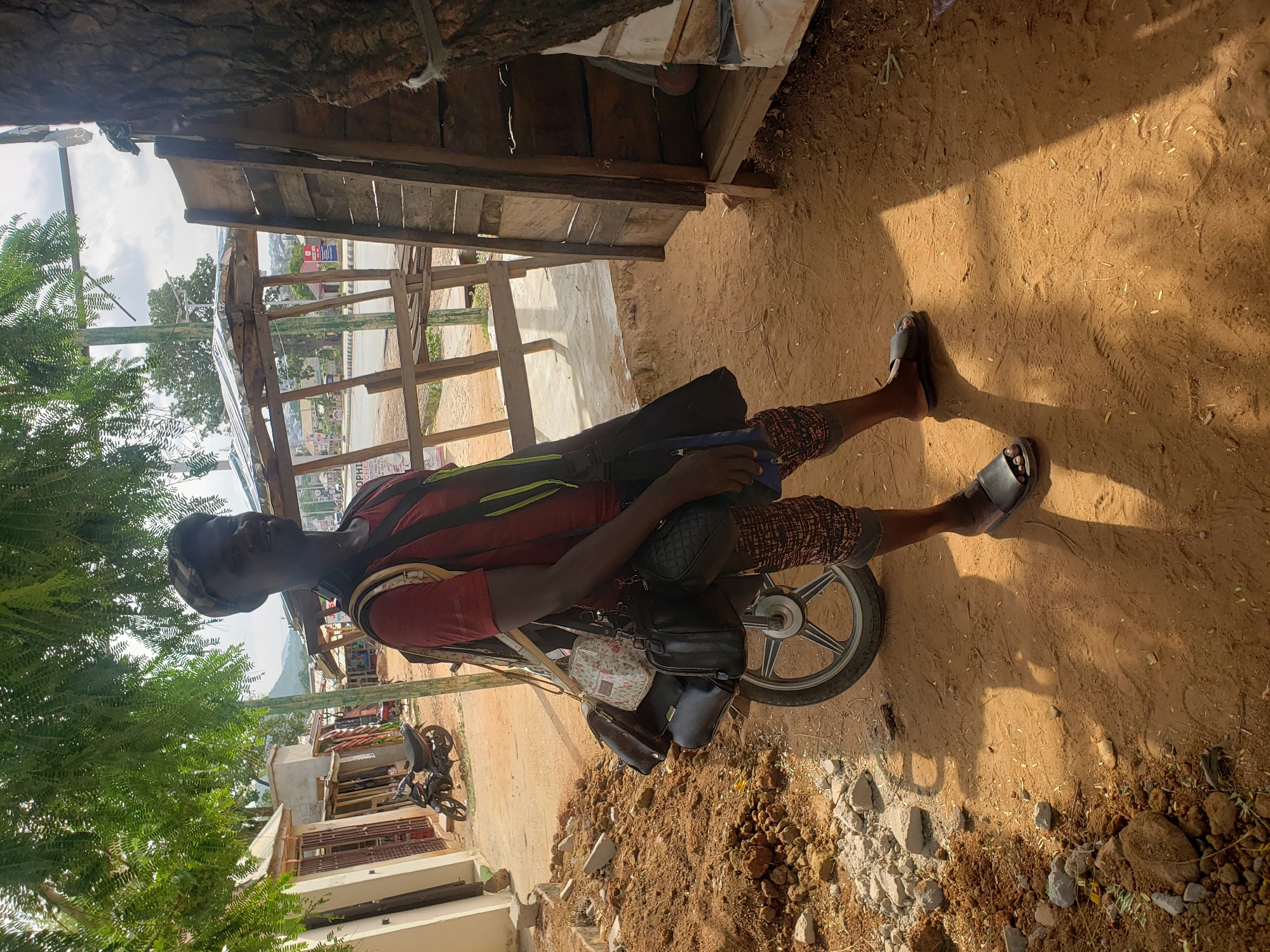 A man stands next to a motorcycle in Adankolo, Nigeria. He is wearing a red t-shirt, patterned shorts, and sandals. He has a bag slung across his chest, and other bags are attached to the motorcycle. The ground is dirt and gravel. A wooden structure provides partial shade, and there are buildings and trees in the background.