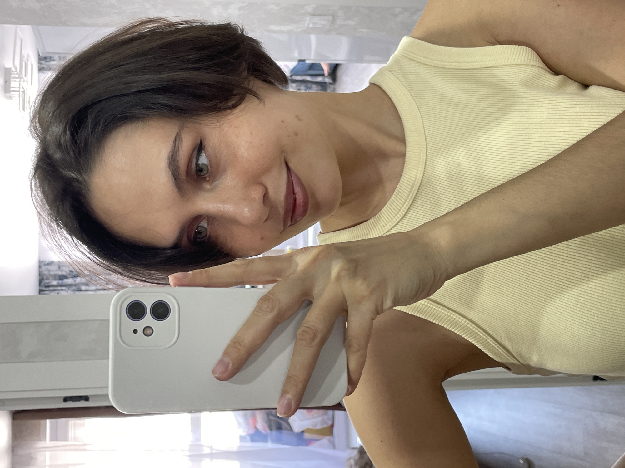 A woman is taking a selfie indoors. She is holding a white smartphone up to her face, and her hand is visible. She has short, dark hair and is wearing a light-yellow tank top. The background appears to be an apartment interior with a window and some furniture. It is impossible to determine if the image was taken in Izhevsk, Russia.